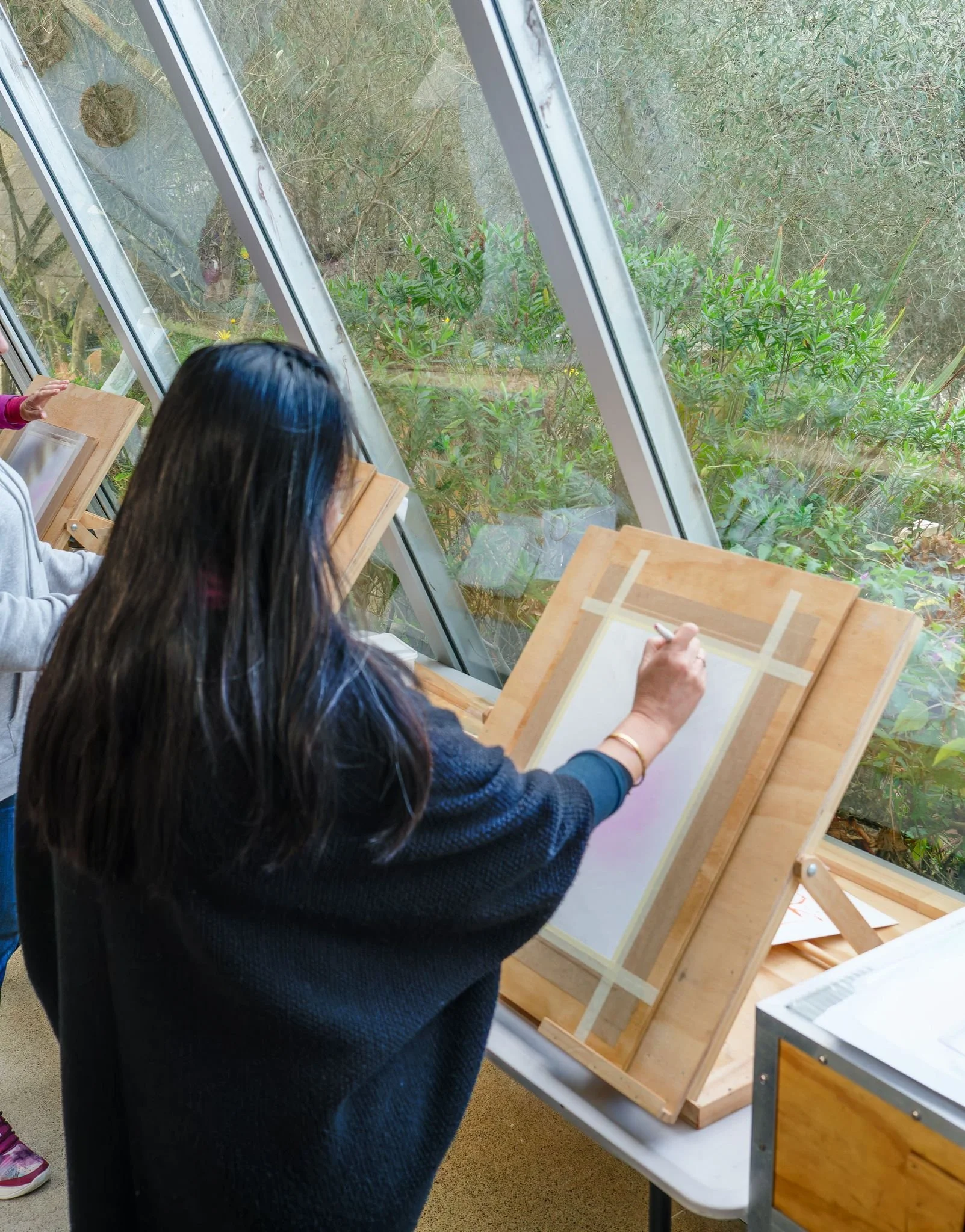 Person sketching on paper at an easel indoors near large windows.