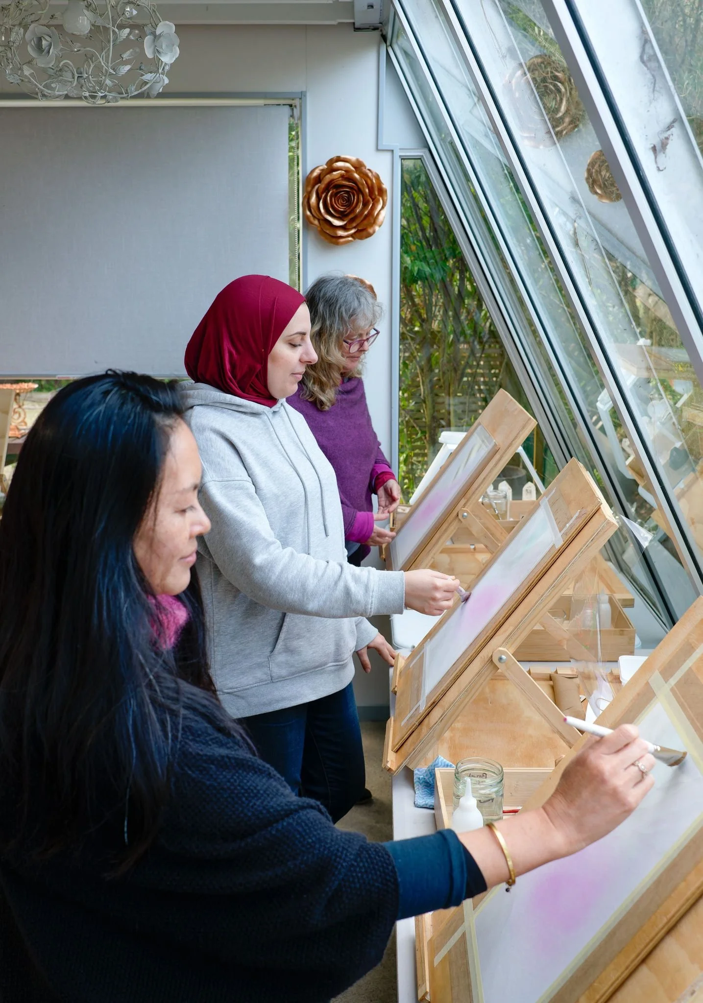 Three women painting at easels indoors next to large windows.