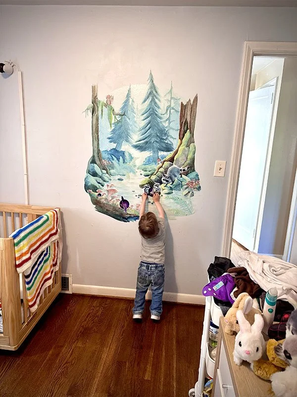 Toddler boy standing on his tippy-toes reaching for the mushroom painted on his peel-and-stick woodland mural. The mural has tall pines, a stream, and a family of raccoons.