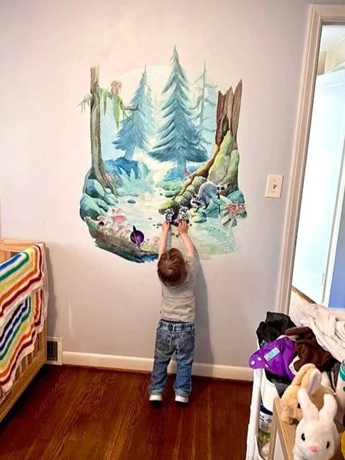 Toddler boy standing on his tippy-toes reaching for the mushroom painted on his peel-and-stick woodland mural. The mural has tall pines, a stream, and a family of raccoons.