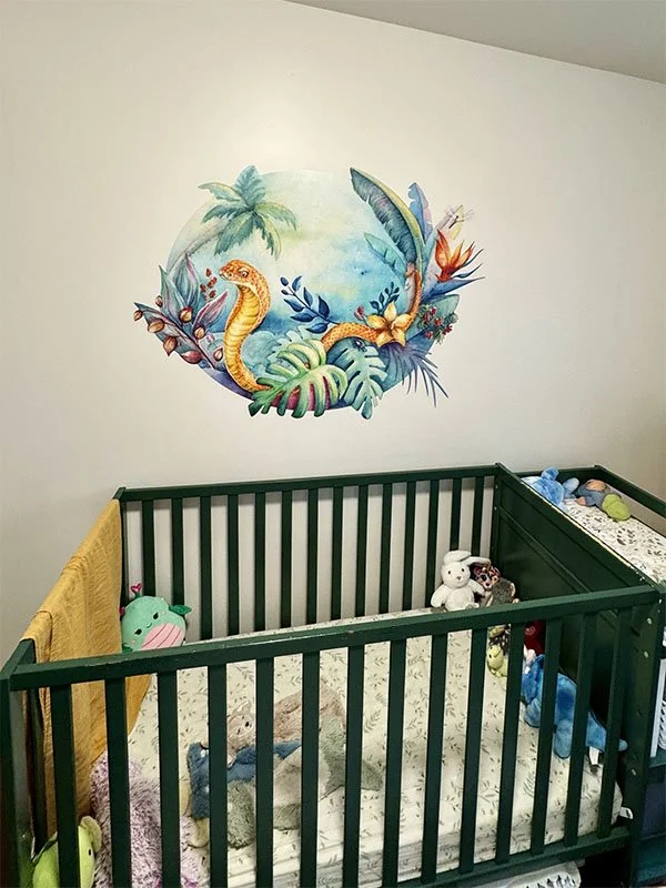 A green baby crib with stuffed animals and toys, a yellow blanket hangs on the side, and a colorful mural of tropical plants and a friendly snake is on the wall behind the crib.