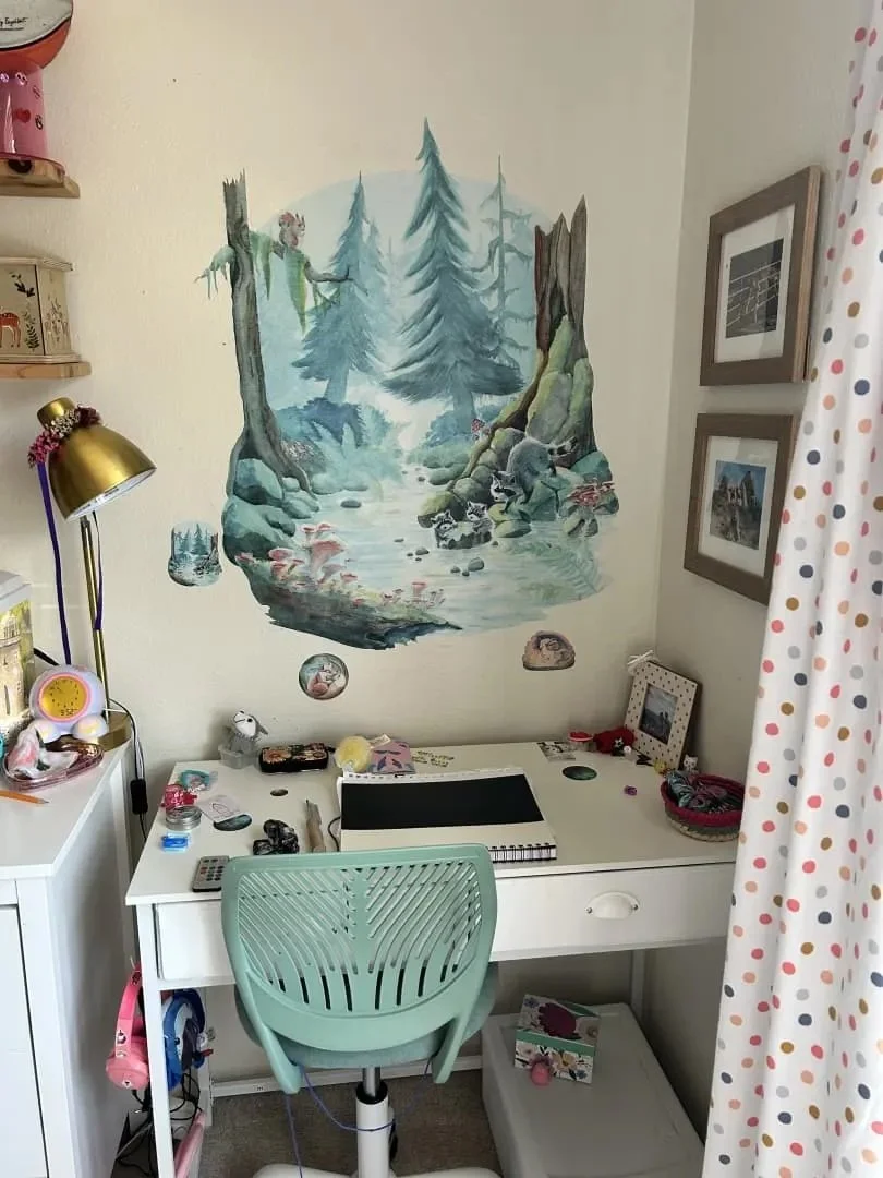 A woodland mural with calming blues and greens is above a white desk. A green chair in front, a white shelf with decorations to the left. The mural shows a mother raccoon leading her kits to down to drink.