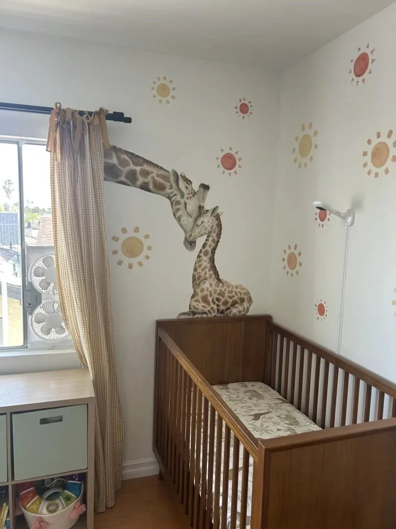 Giraffe mural above a crib with a window and curtains in a safari-themed nursery. The mother giraffe's head is reaching down through the window and caressing her calf.