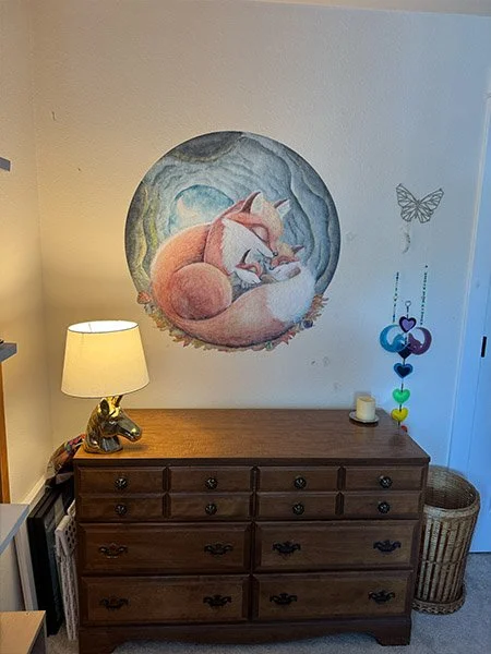 Wooden dresser in a child's room with a wicker basket. Hand-painted mural of a mother fox curled up with her two cubs on the wall.