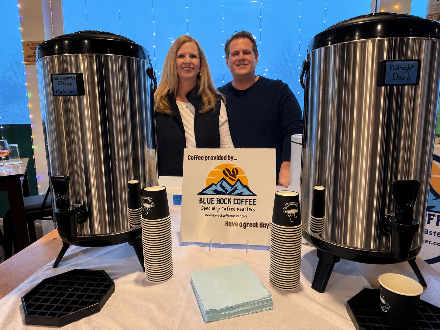 Sue and John Krepelka are smiling as they stand behind a coffee station with two large coffee dispensers. There are paper cups stacked and a sign that reads 'Coffee provided by Blue Rock Coffee, Specialty Roasters. Have a great day!'