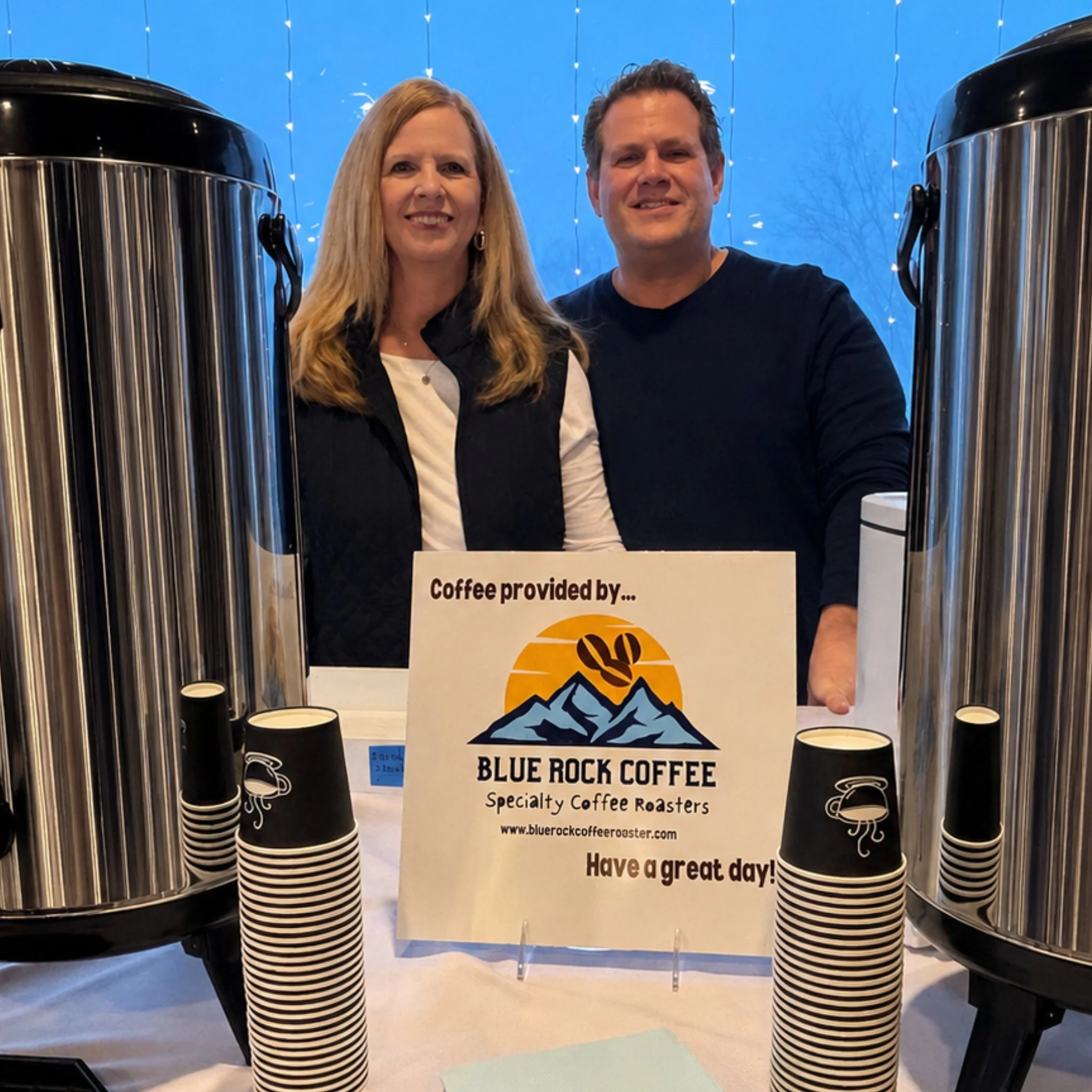 Sue and John Krepelka are smiling as they stand being a table with coffee cups and a sign that reads "Coffee provided by Blue Rock Coffee, Specialty Coffee Roasters. Have a great day!"