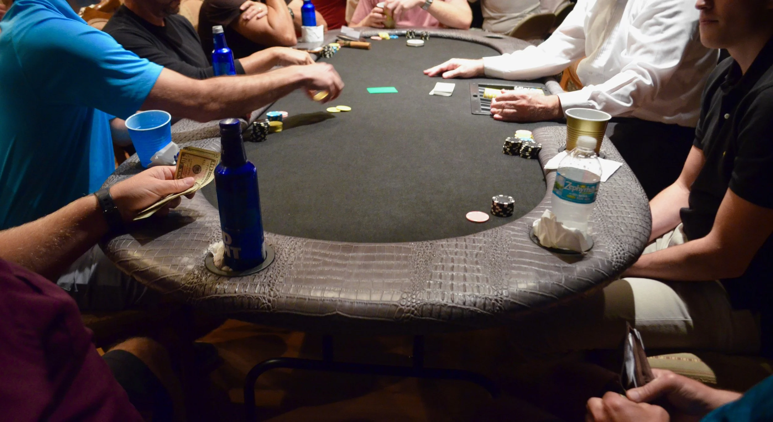 A group of people sitting around a poker table playing cards and betting chips, with drinks and water bottles on the table.