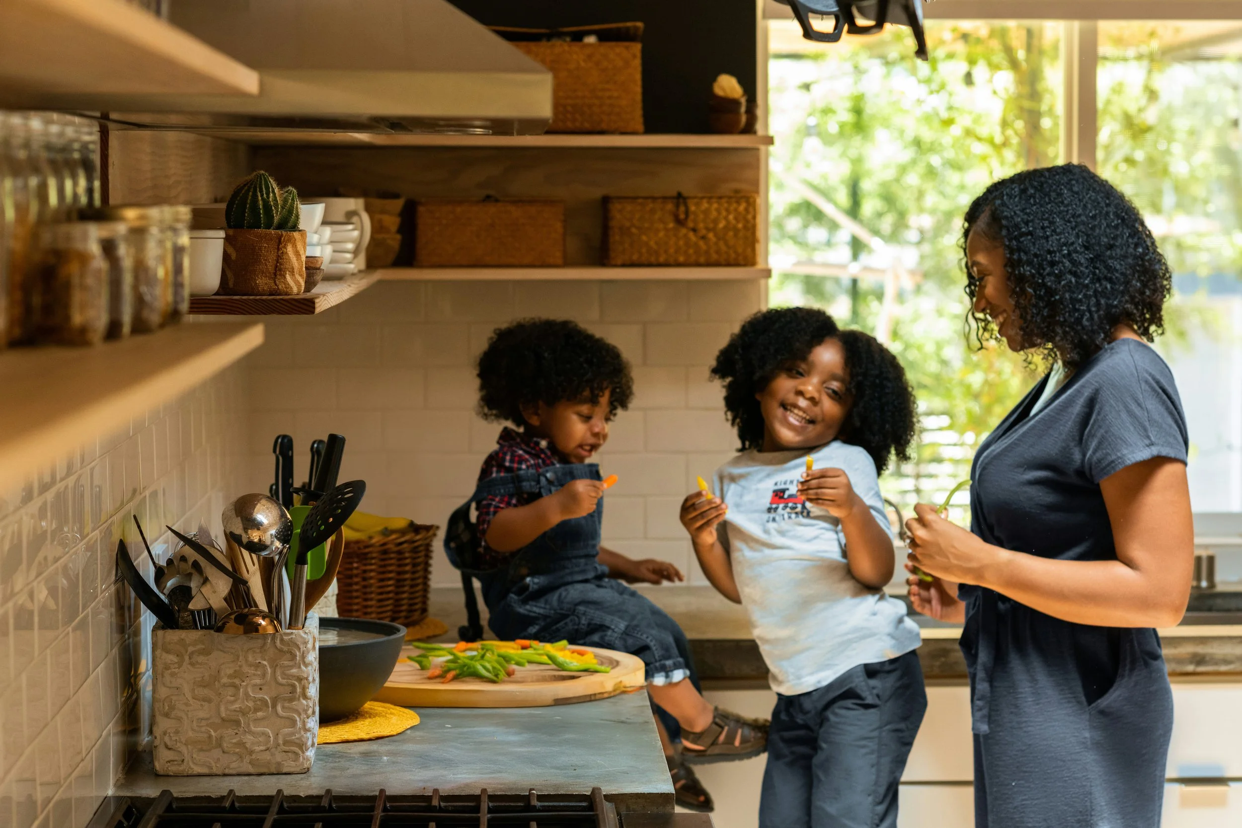 A woman and two children in a kitchen with sunlight coming through the window, preparing vegetables on a countertop.
