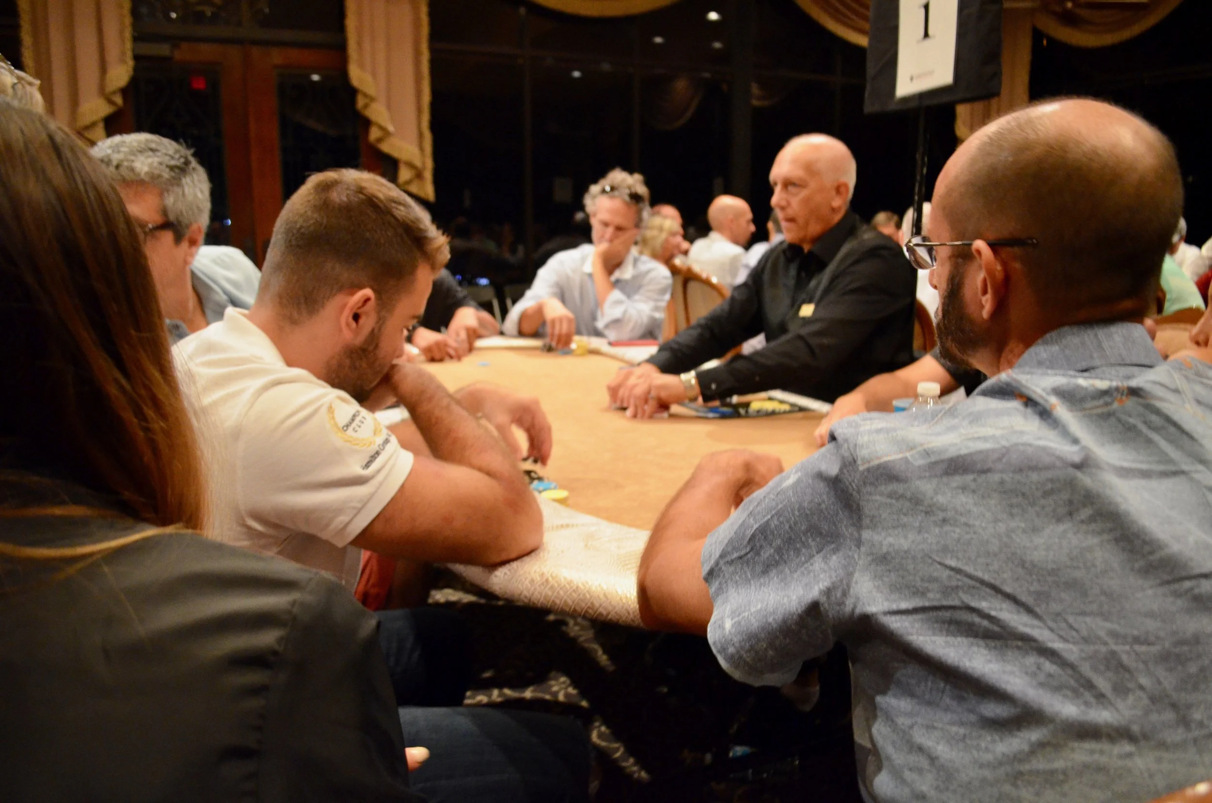 People sitting around a poker table in a dimly lit room, playing poker, with some smiling and others focused on the game.