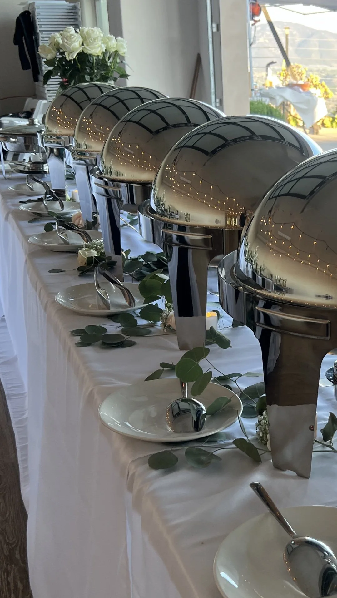 Buffet setup with covered chafing dishes, decorative plates, silverware, and white roses on a table.