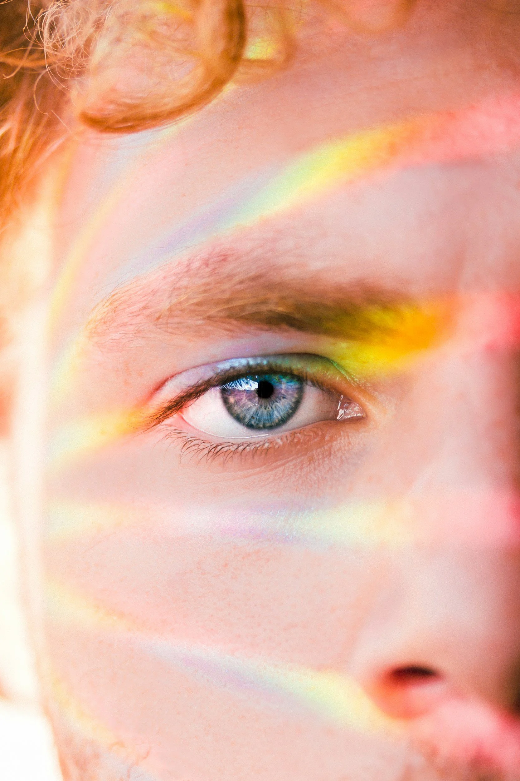 Close-up of a young person's face, focusing on their blue eye with rainbow light reflections on their skin.