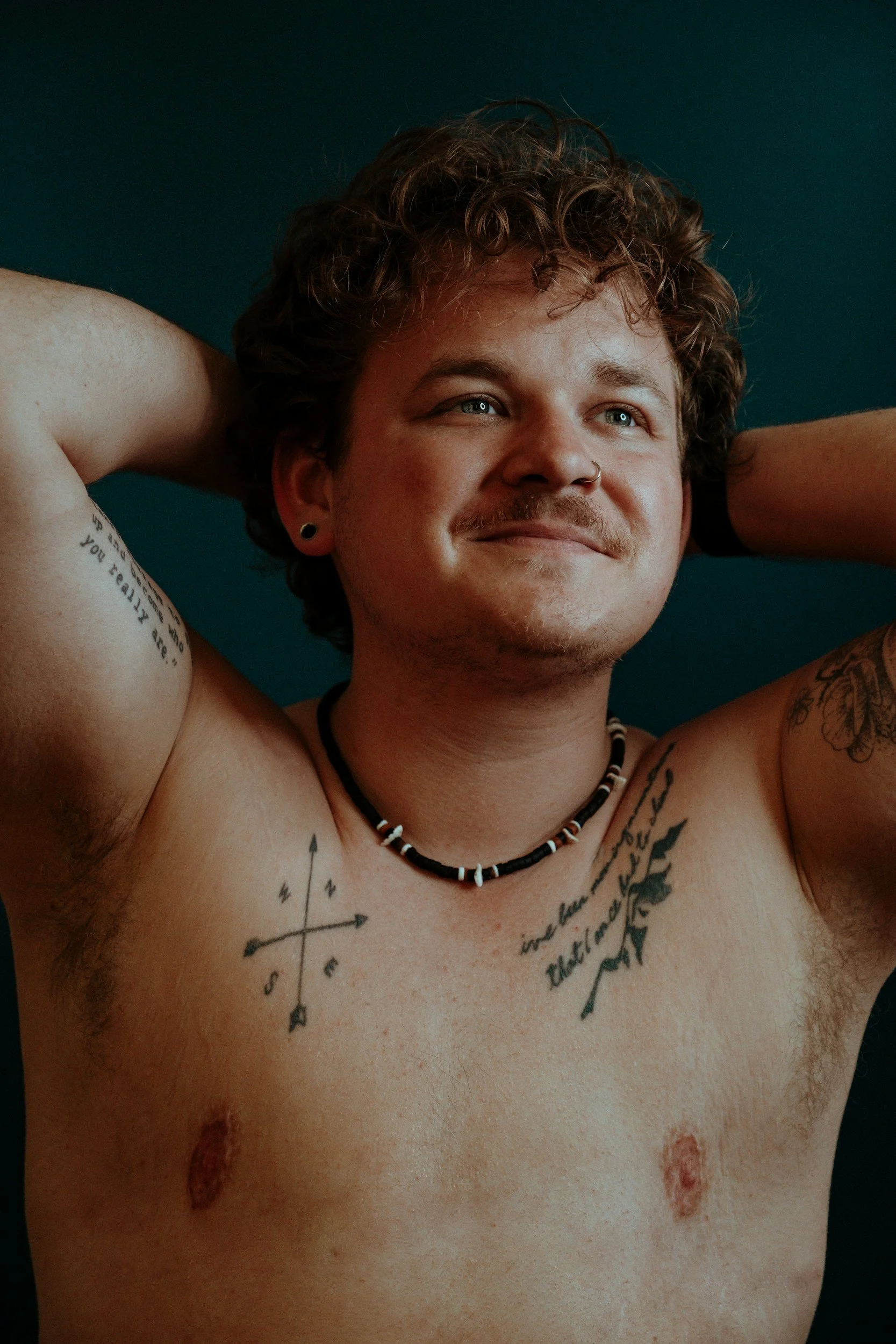 A shirtless young man with curly hair, blue eyes, and a nose piercing lying down against a dark background. Looking for support with his gender affirming care and booking a session with Grey Insight.