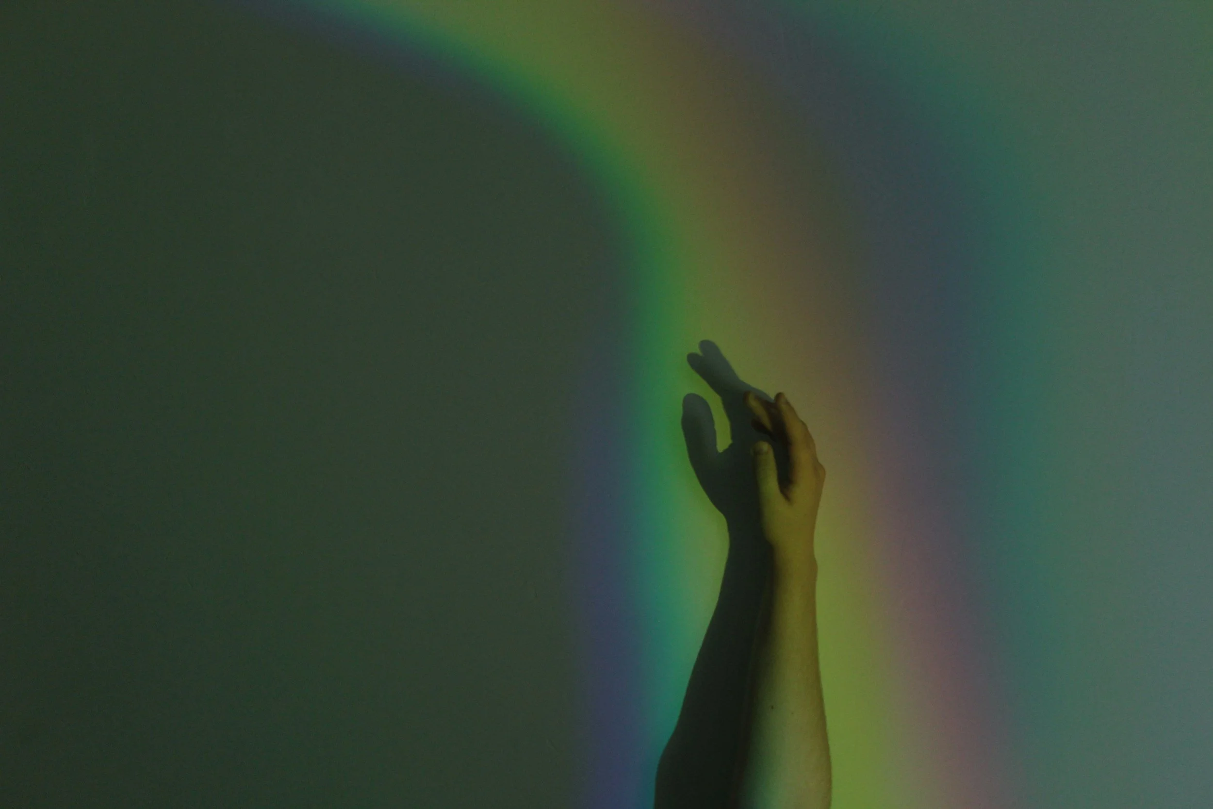 A hand reaching out towards a colorful, blurred background with shades of green, yellow, and blue. Looking for support with their gender affirming care with Grey Insight