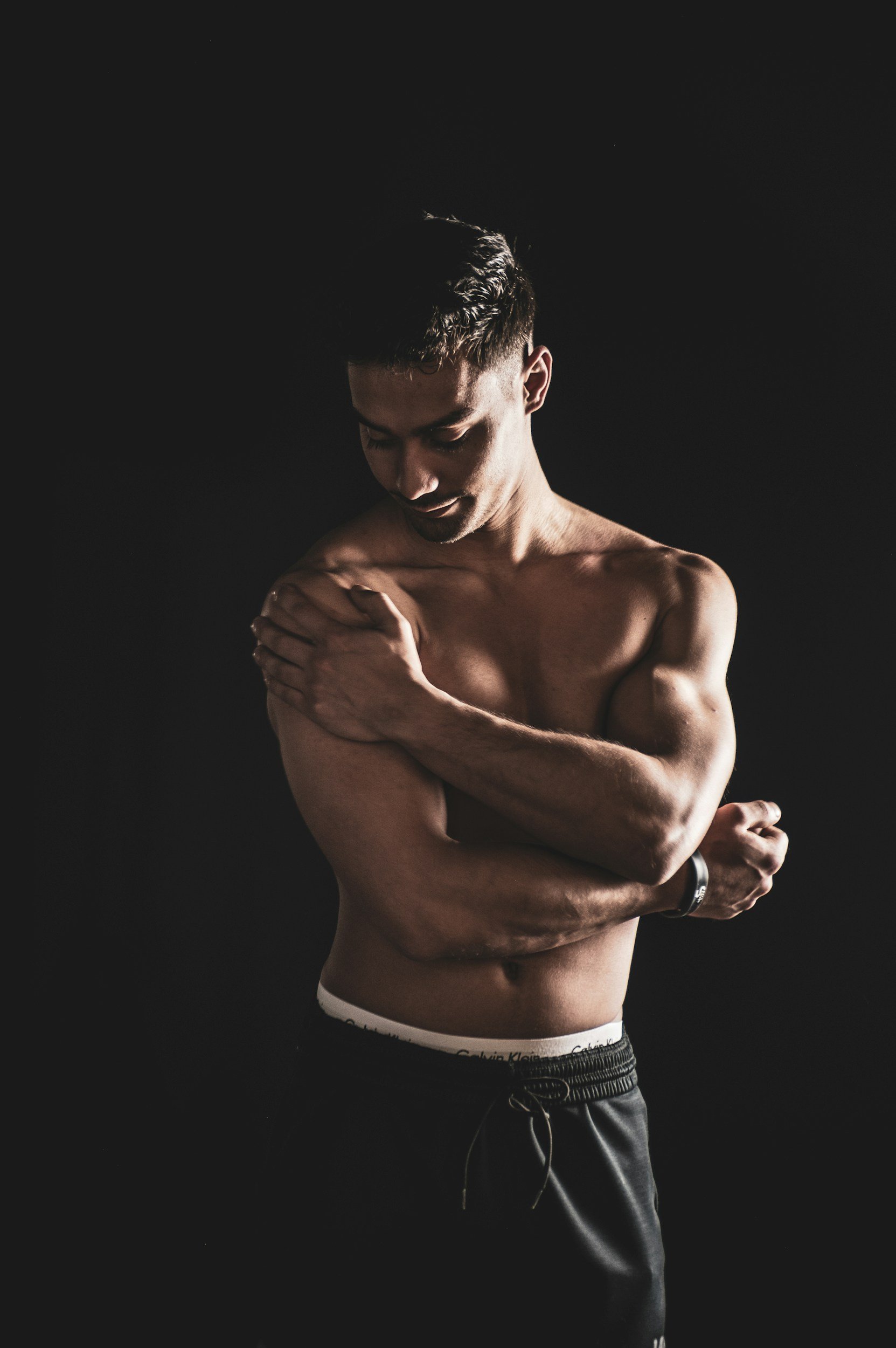 A shirtless man with short dark hair and a muscular build, standing against a black background, looking down, with his arms crossed over his chest and his right hand touching his left shoulder.