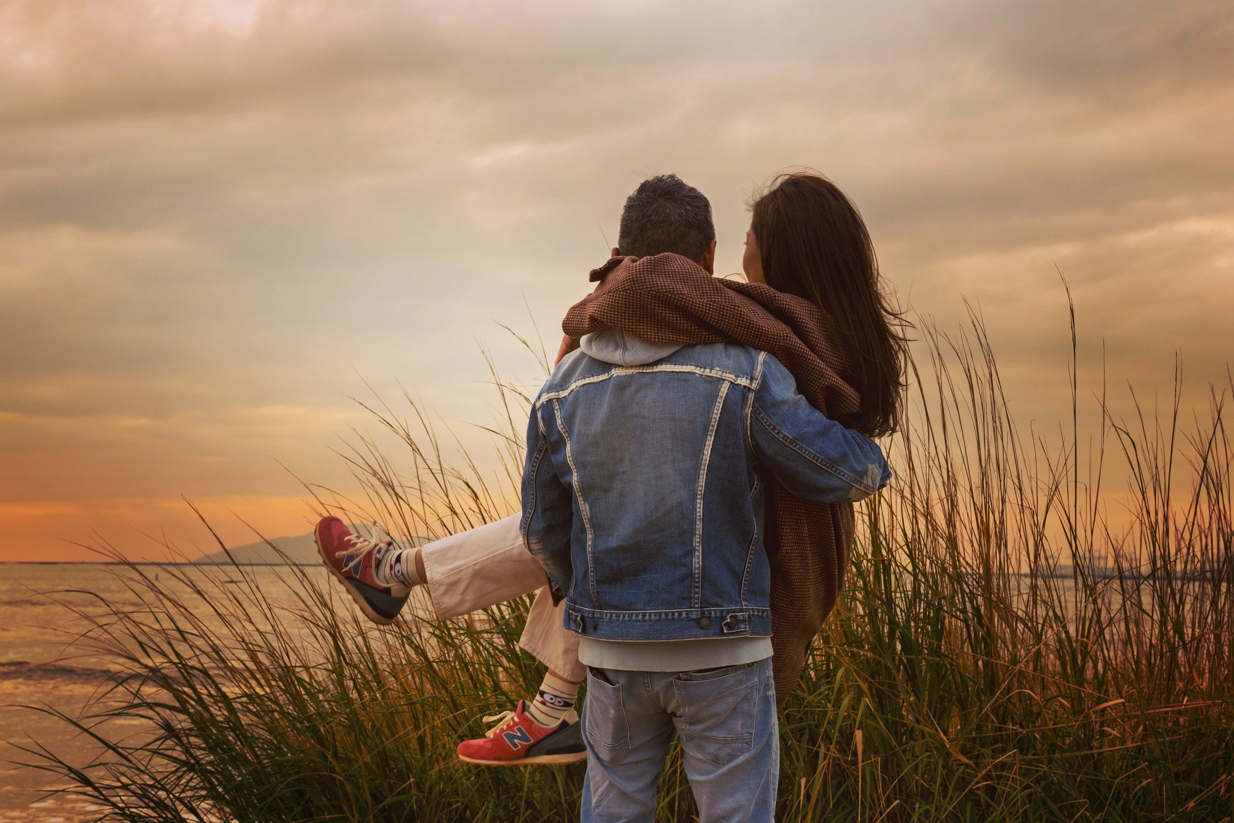 A couple sitting near the water during sunset, with the man holding the woman on his lap. The woman is leaning into him, and they are both looking at the sunset sky. The scene is surrounded by tall grasses.