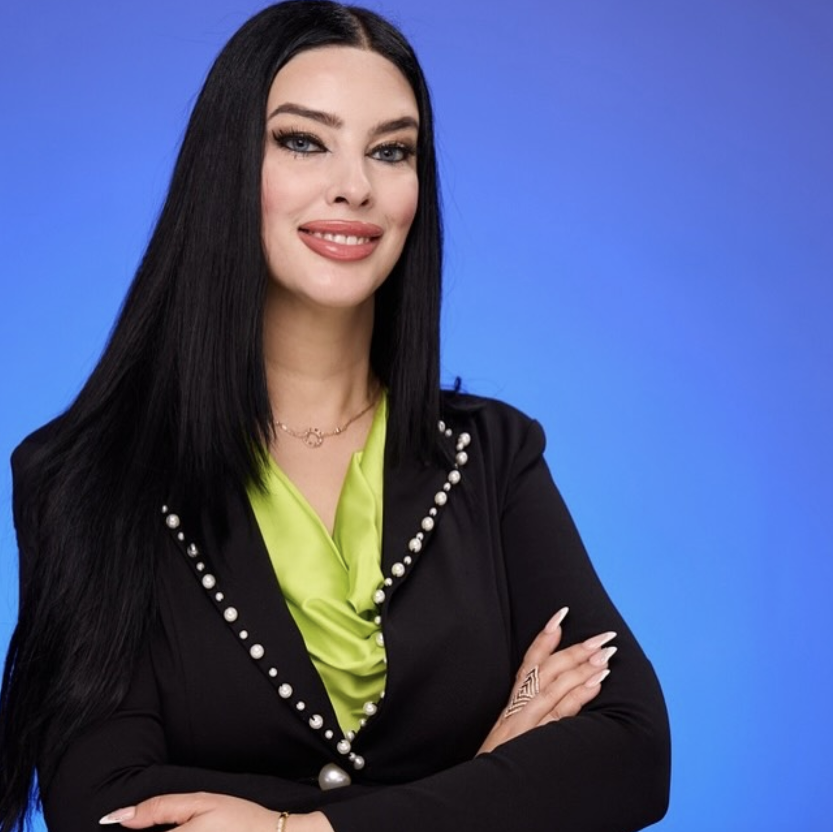 A woman with long black hair, blue eyes, and makeup, wearing a black blazer with pearl embellishments, a neon green blouse, and jewelry, standing against a blue background.