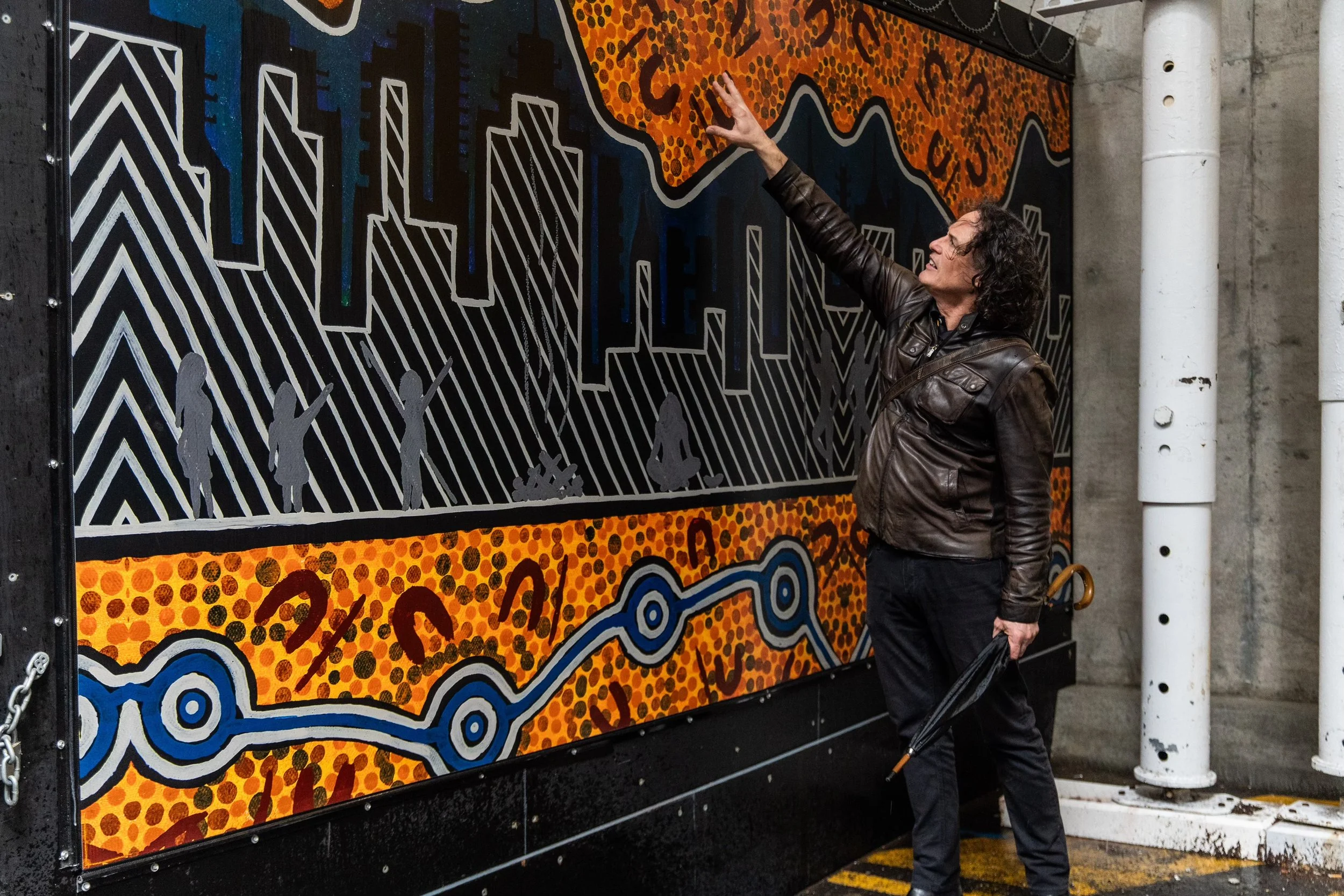 A man in a brown leather jacket and black pants pointing at a large, colorful mural that features abstract and cityscape elements, alongside figures of children and desert animals.