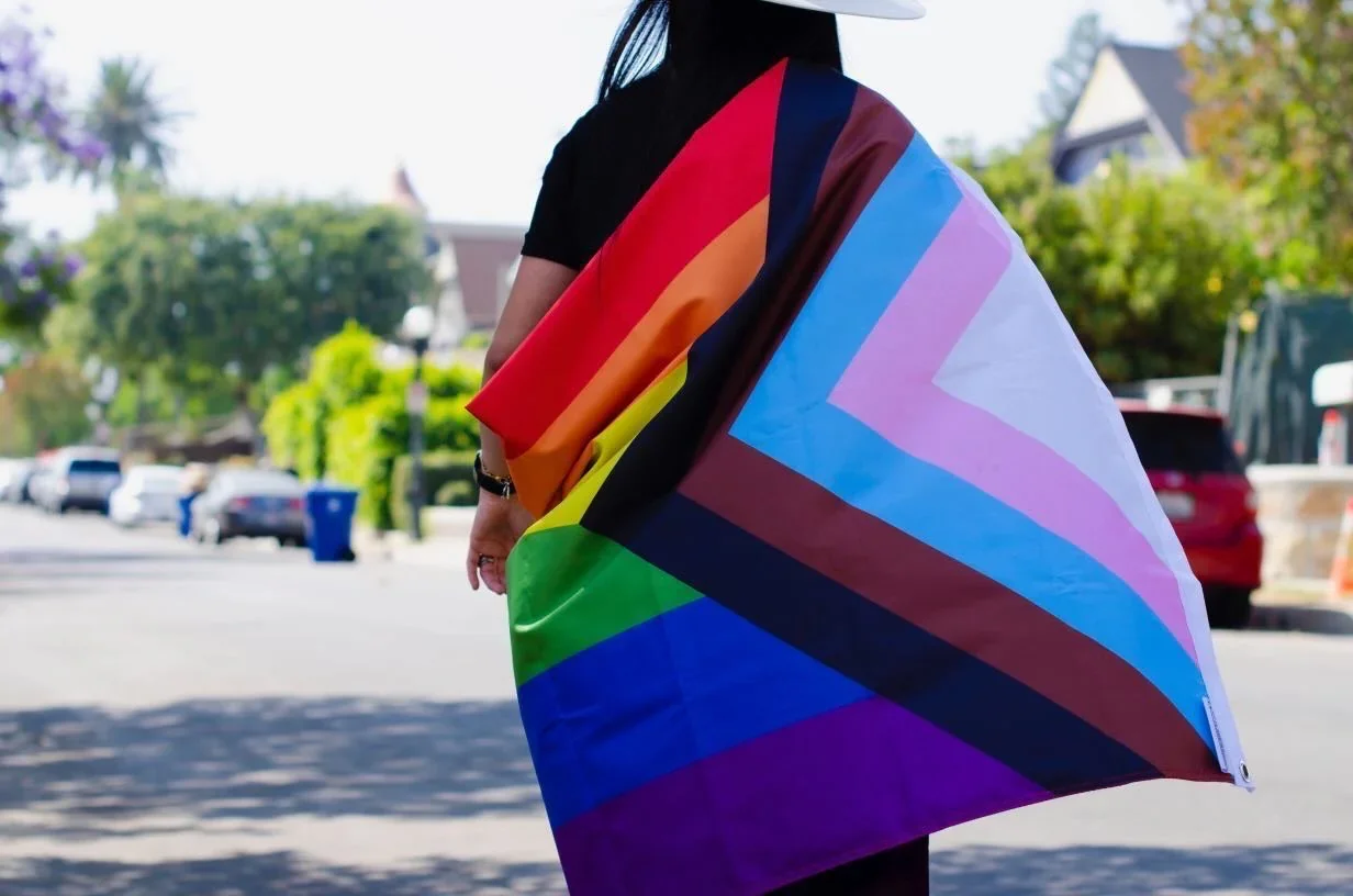 Person holding an LGBTQ+ pride flag with rainbow colors, walking outdoors in a neighborhood with trees and houses in the background.