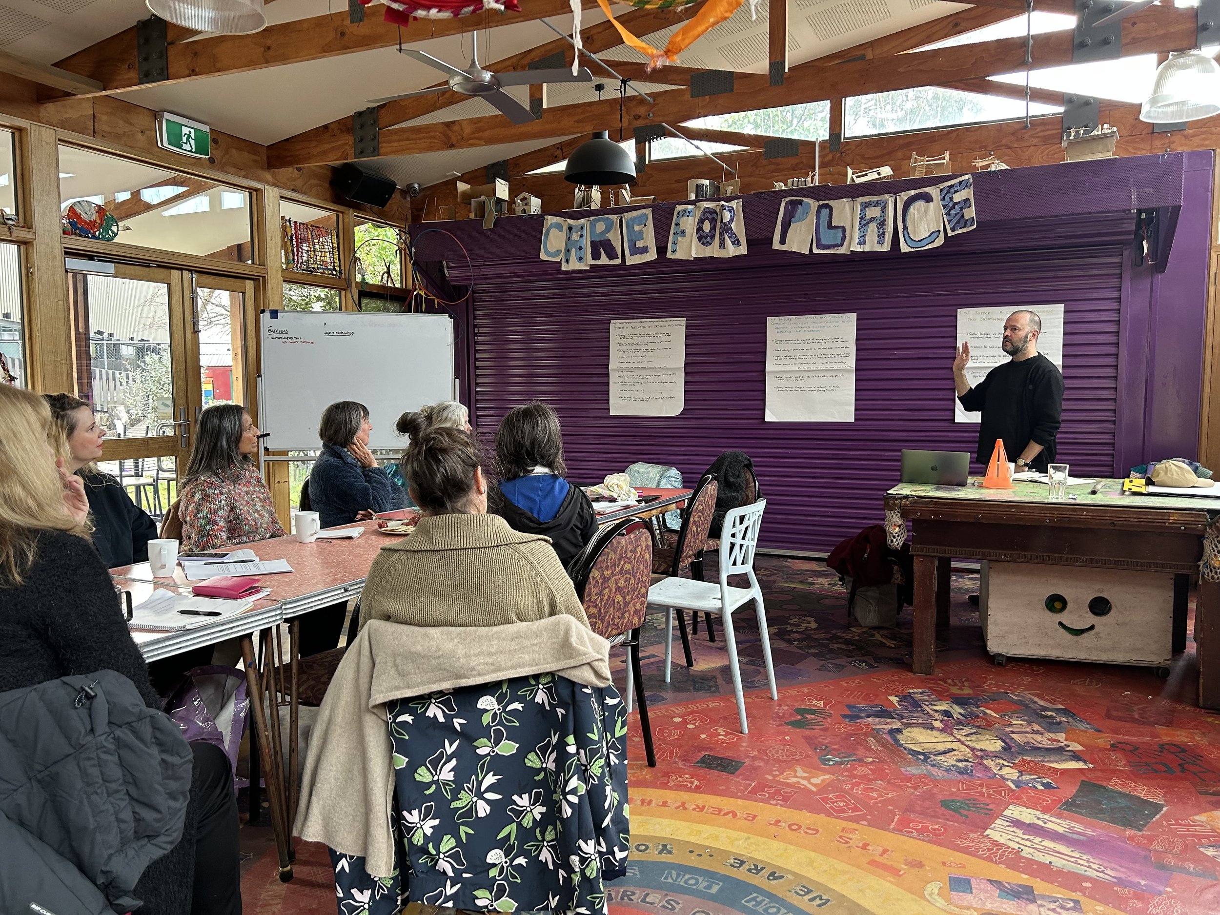 An indoor workshop or seminar with a man giving a presentation to an audience of women seated at tables. The room has wooden beams, a whiteboard, and a purple backdrop with a banner that reads 'CARE FOR PLACE.'