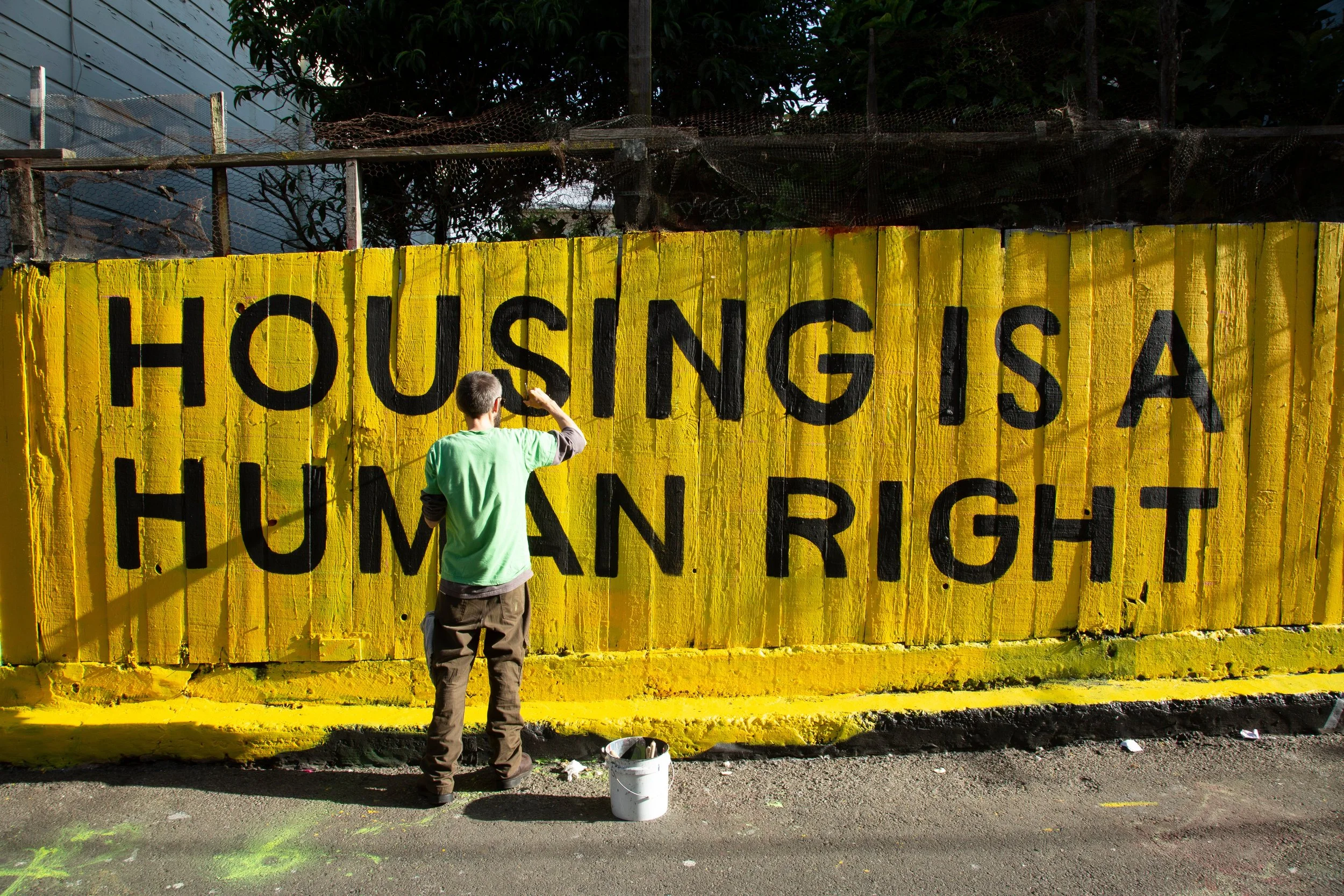 A person painting a mural on a yellow wall that reads, 'Housing is a Human Right'