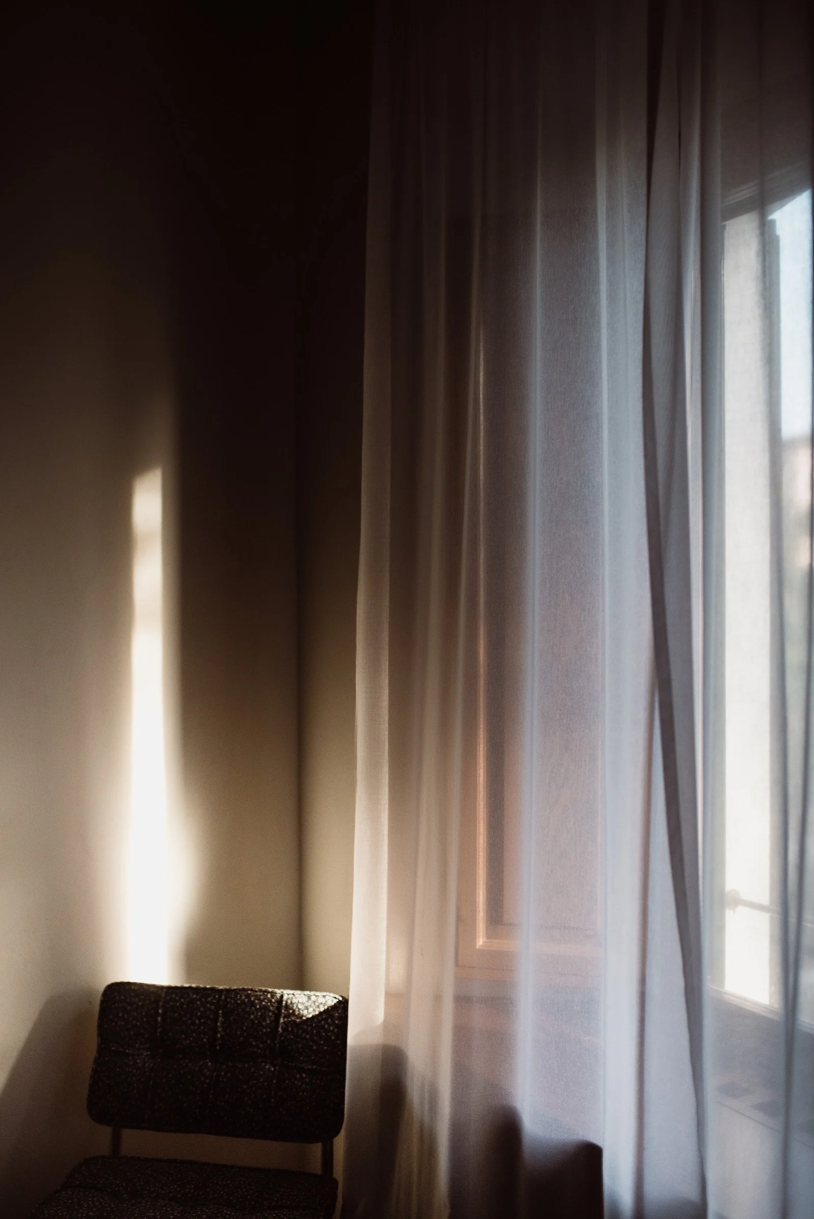 Sunlight streaming through sheer curtains onto a chair placed against a wall in a psychologist's therapy room with a window.