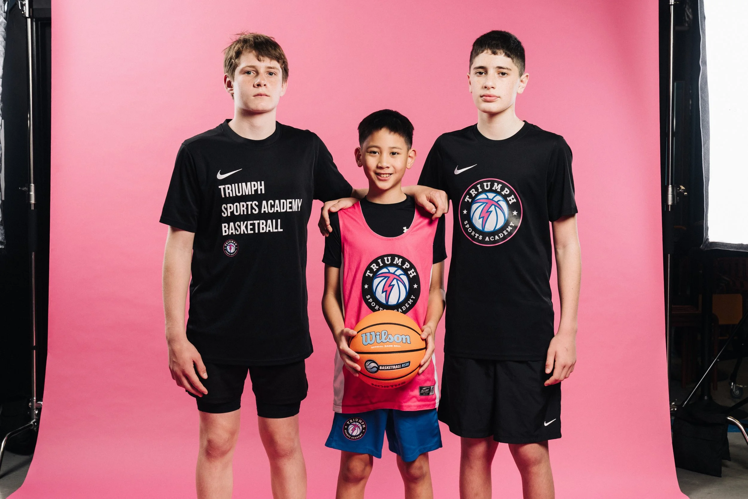 Three boys in sports attire posing for a photo on a pink backdrop. The boy in the middle is holding a basketball and wearing a pink and black jersey. The two taller boys on either side are wearing black jerseys with sports logos.