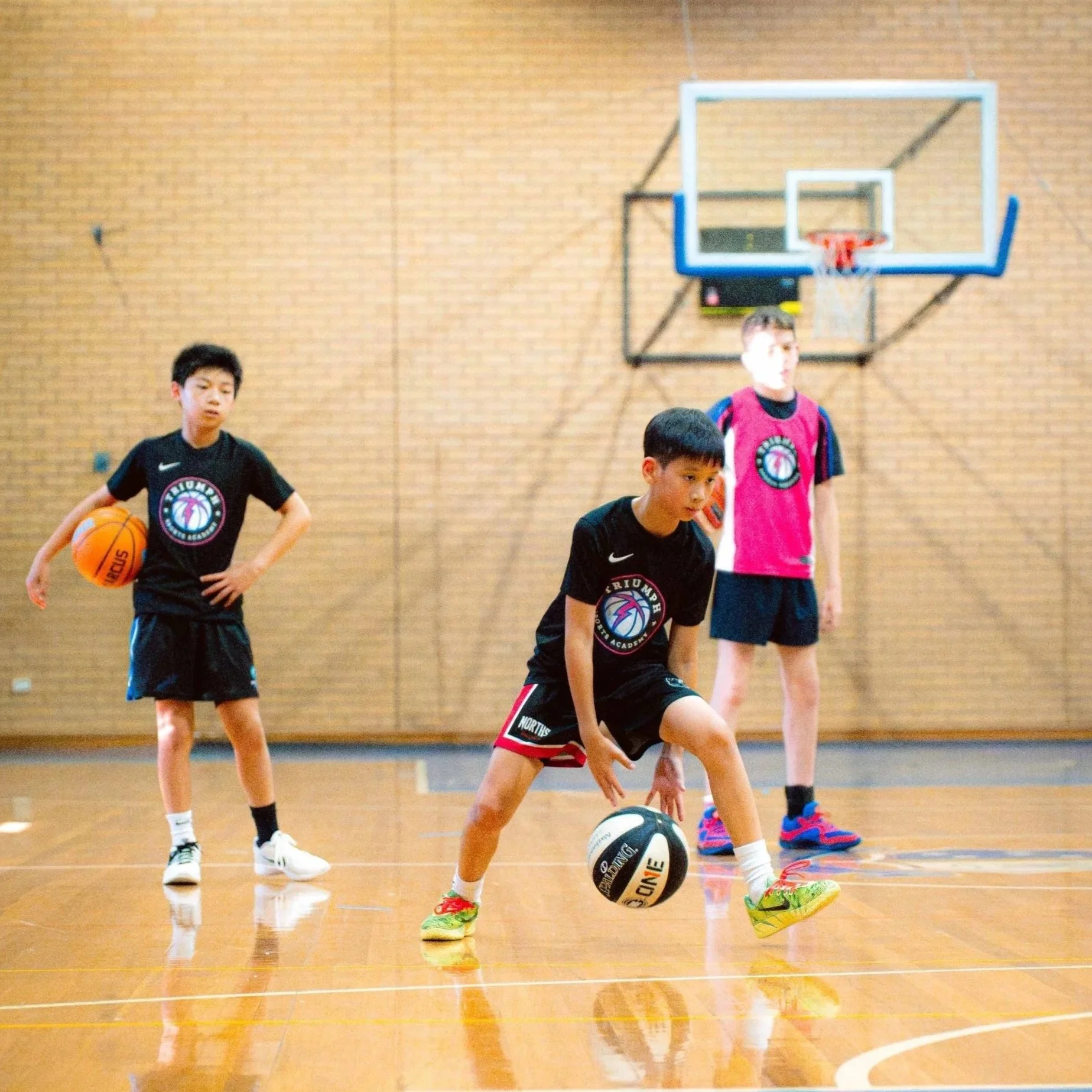 Young boys playing basketball in an indoor gymnasium with three boys visible, one actively shooting or practicing with a basketball, while the others stand nearby