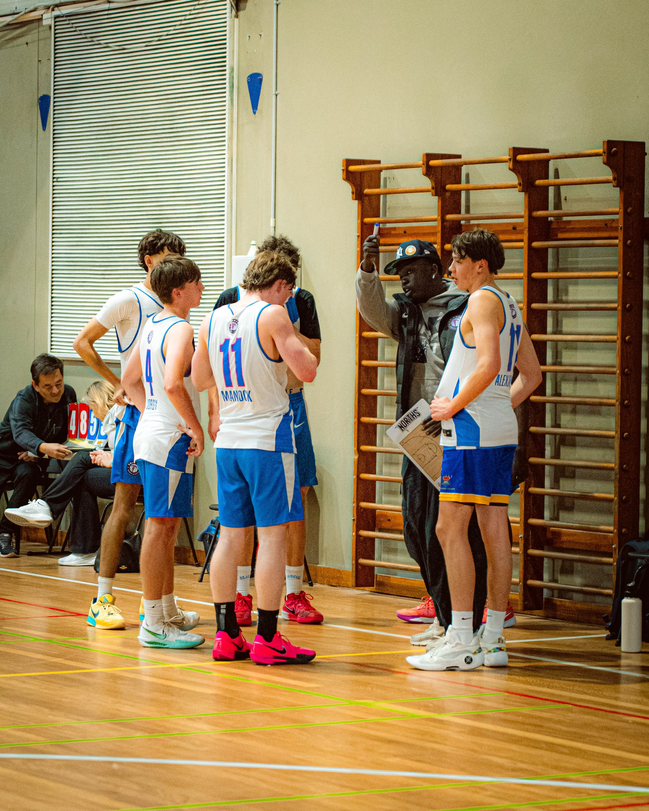 A basketball coach is talking to young players during a timeout in a gymnasium. The players are wearing white and blue uniforms. There are others seated and standing nearby, and basketball equipment is visible.