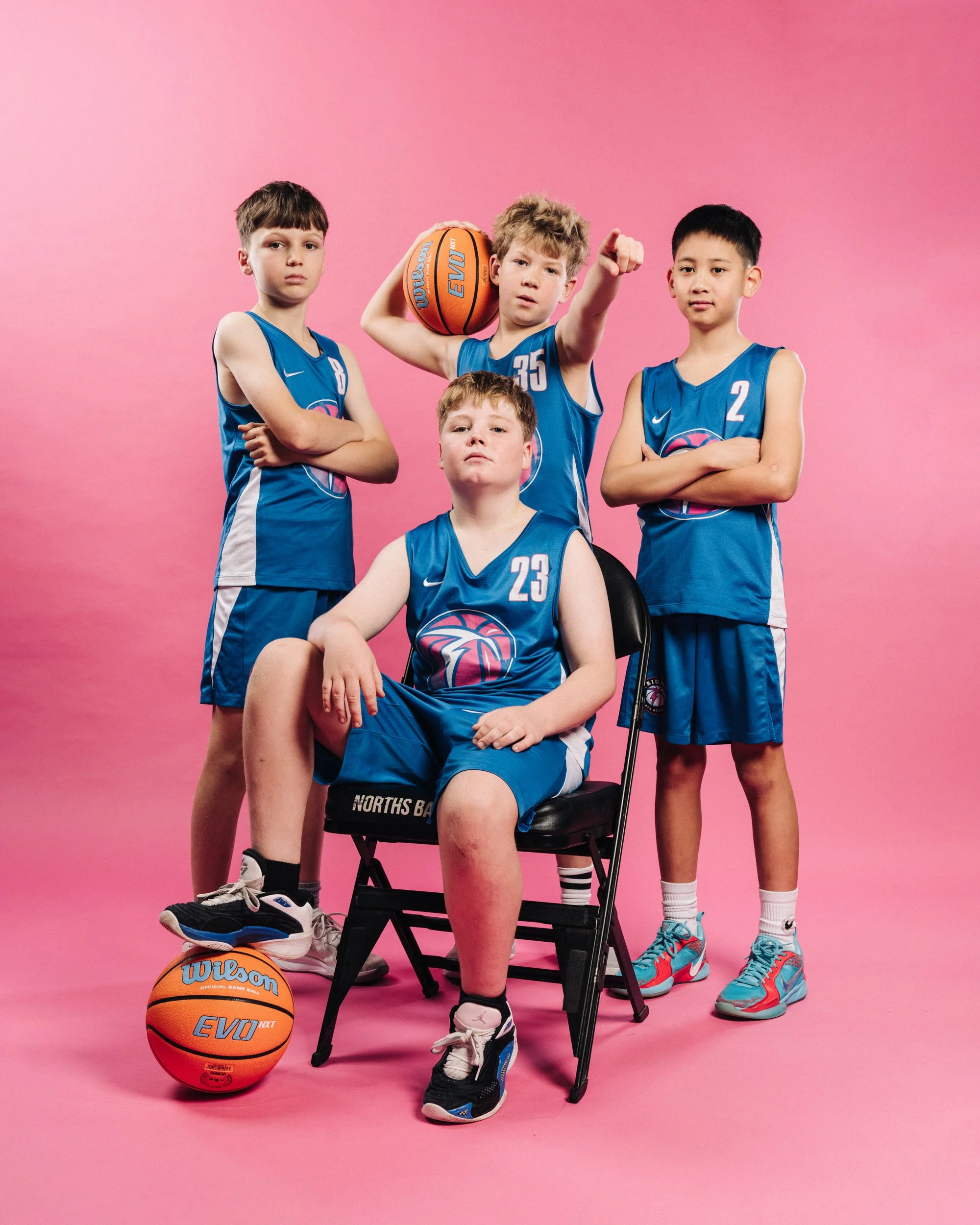 A boys' youth basketball team dressed in blue basketball uniforms, posing against a pink background. One player is seated in a chair with a knee injury, and the other four players stand around him.