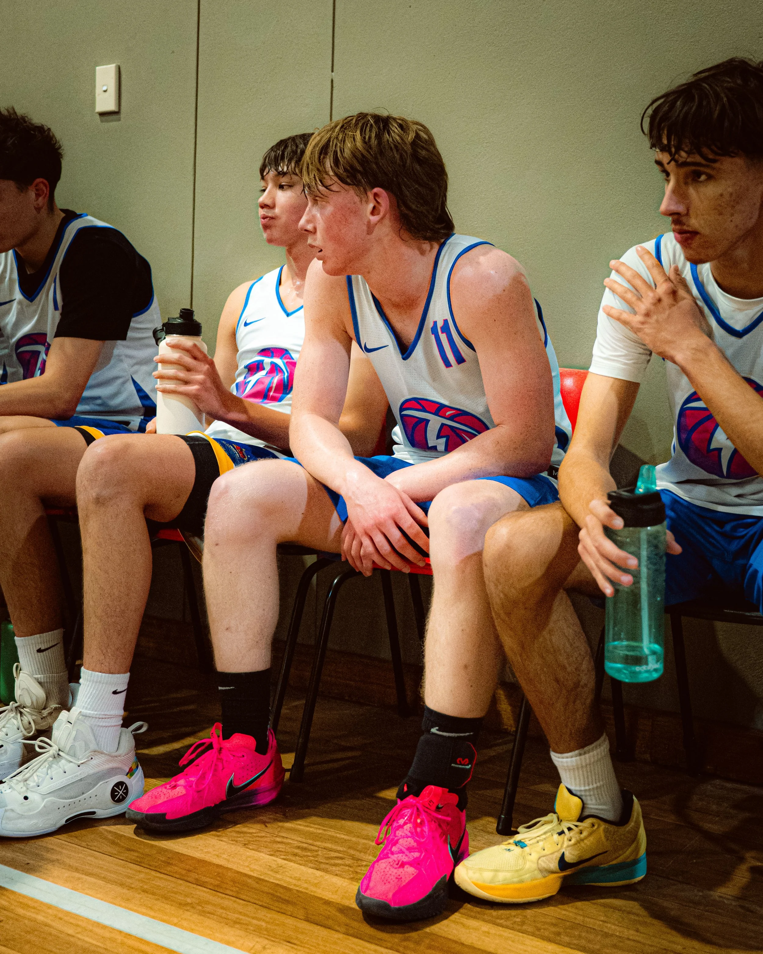 Young male basketball players sitting on a bench during a game or practice, wearing white jerseys with blue accents and holding water bottles.