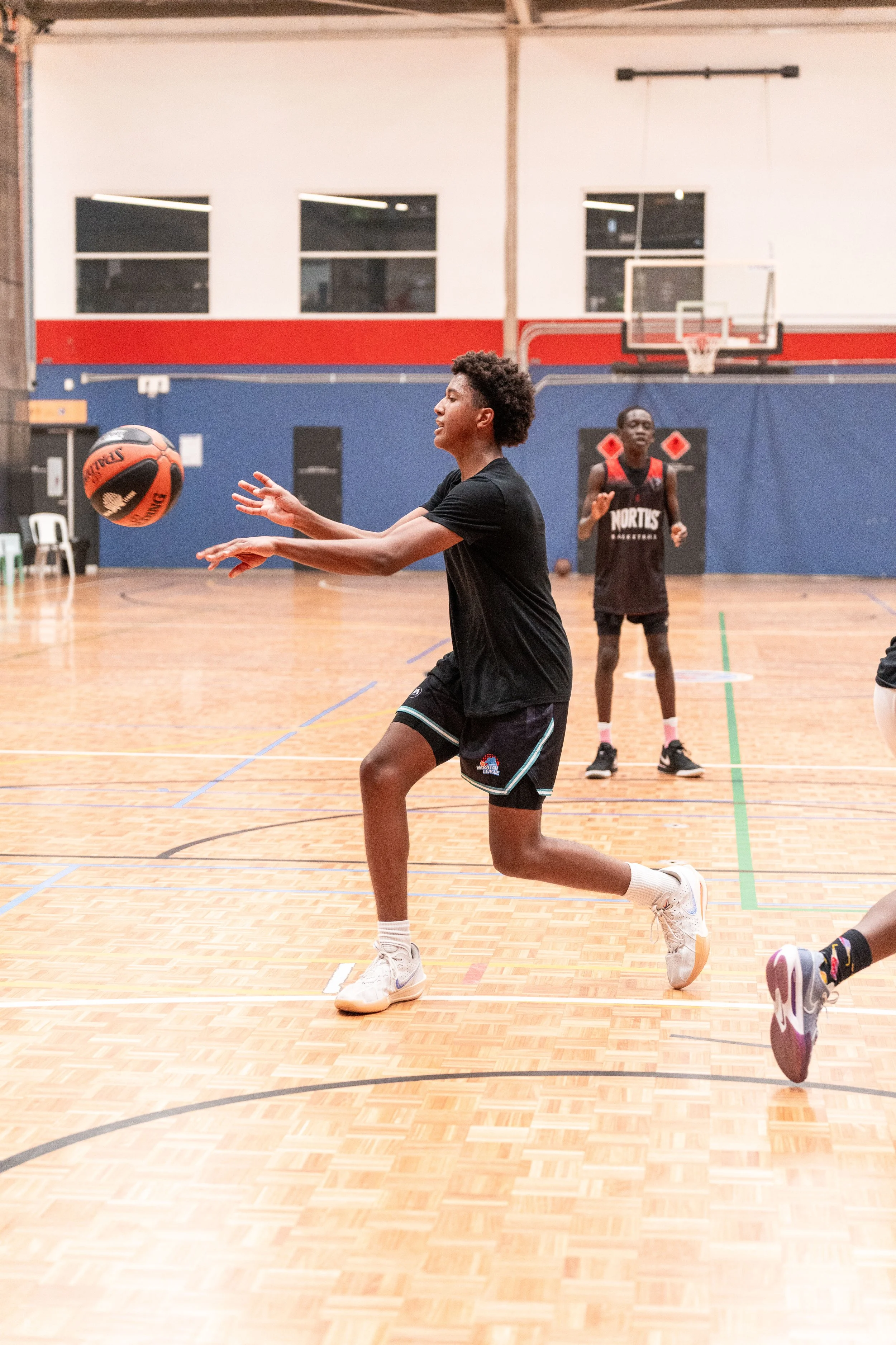 A young man with dreadlocks holding a basketball in a gymnasium, looking attentively to the side, wearing a white sports jersey with a logo on it.