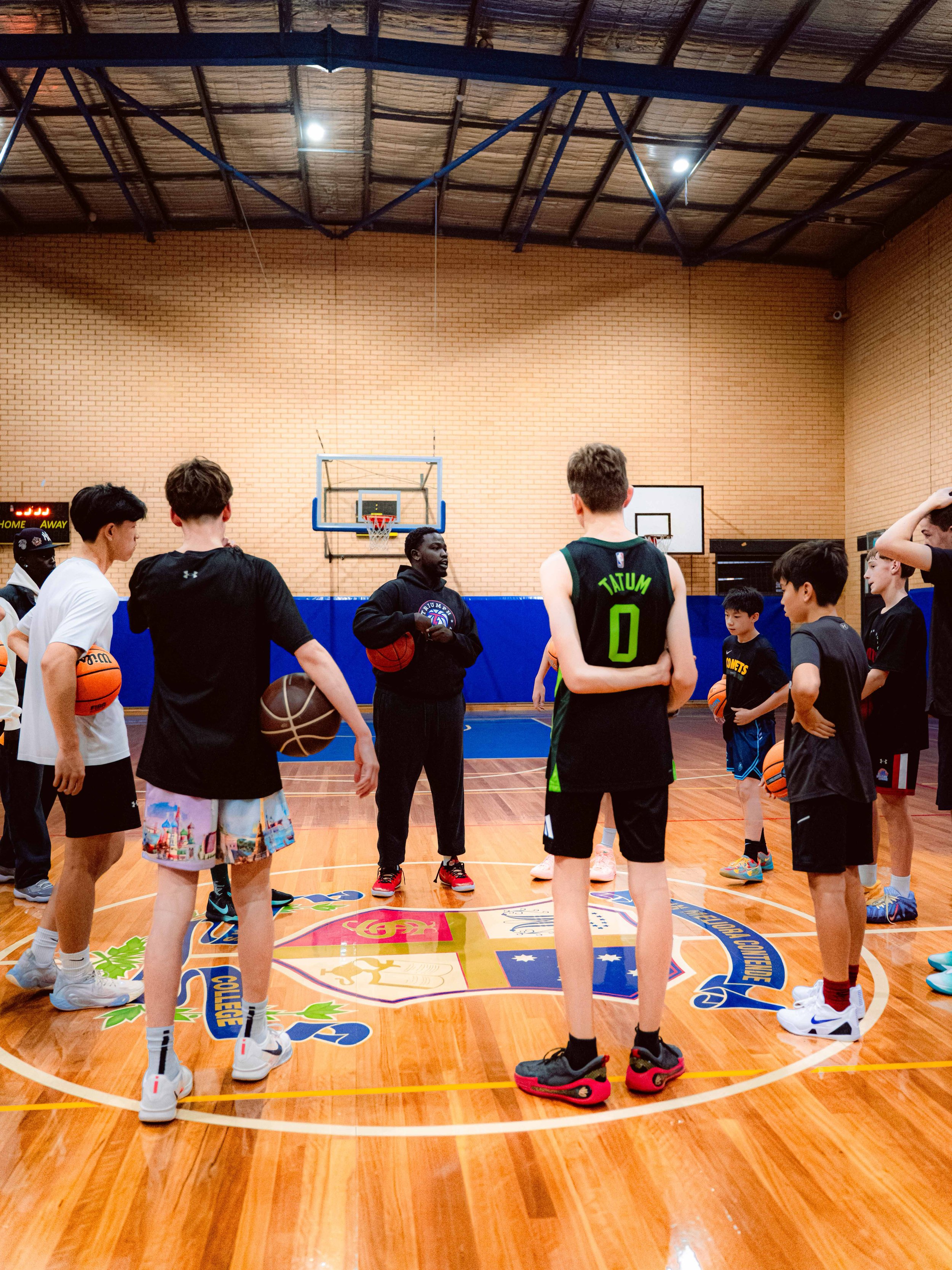 A young man wearing a white sports shirt, black shorts, and a black headband holding a red and white basketball on an indoor court, with two people blurred in the background.