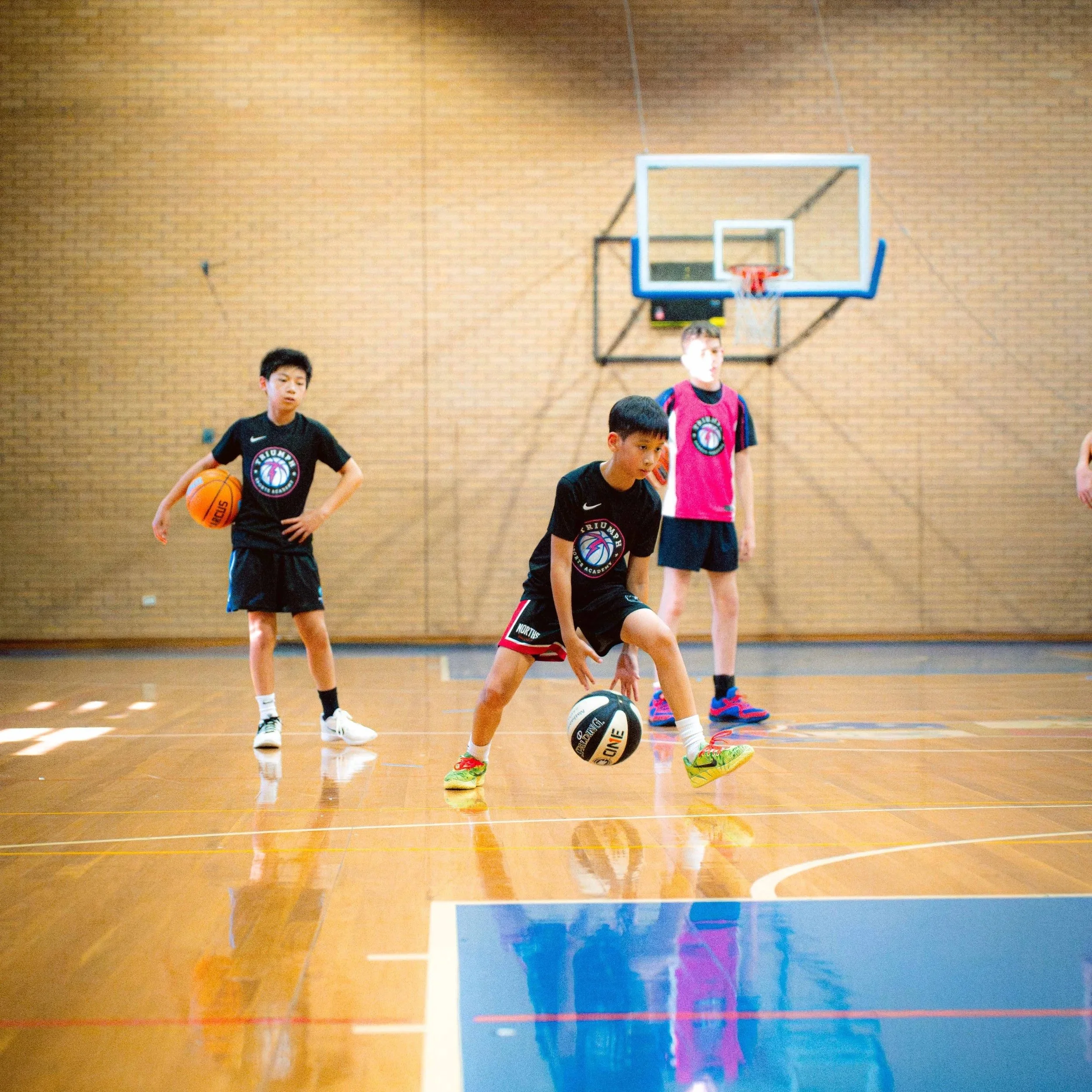 Three boys playing basketball in an indoor gym. One boy is dribbling a basketball, another holding a ball, and the third standing near the basket. All are wearing sports uniforms.