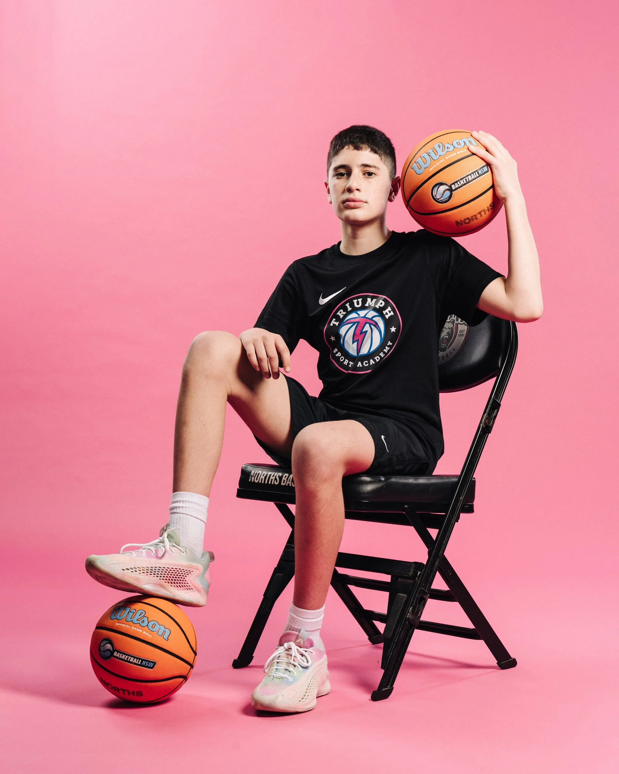Young basketball player sitting on a black chair with a pink background, holding a basketball on his shoulder, with another basketball under his foot, wearing a black t-shirt and shorts, white sneakers, and pink socks.