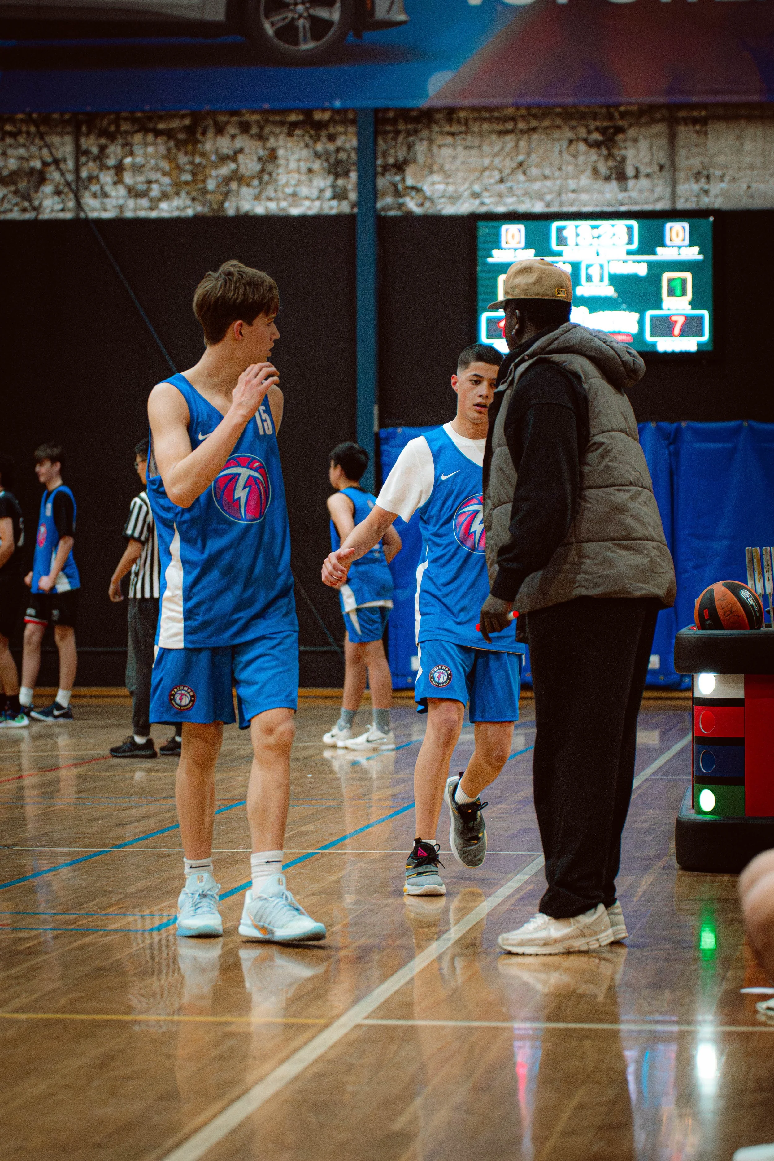 A youth basketball coach speaks to two players in blue uniforms during a game or practice in an indoor gymnasium.