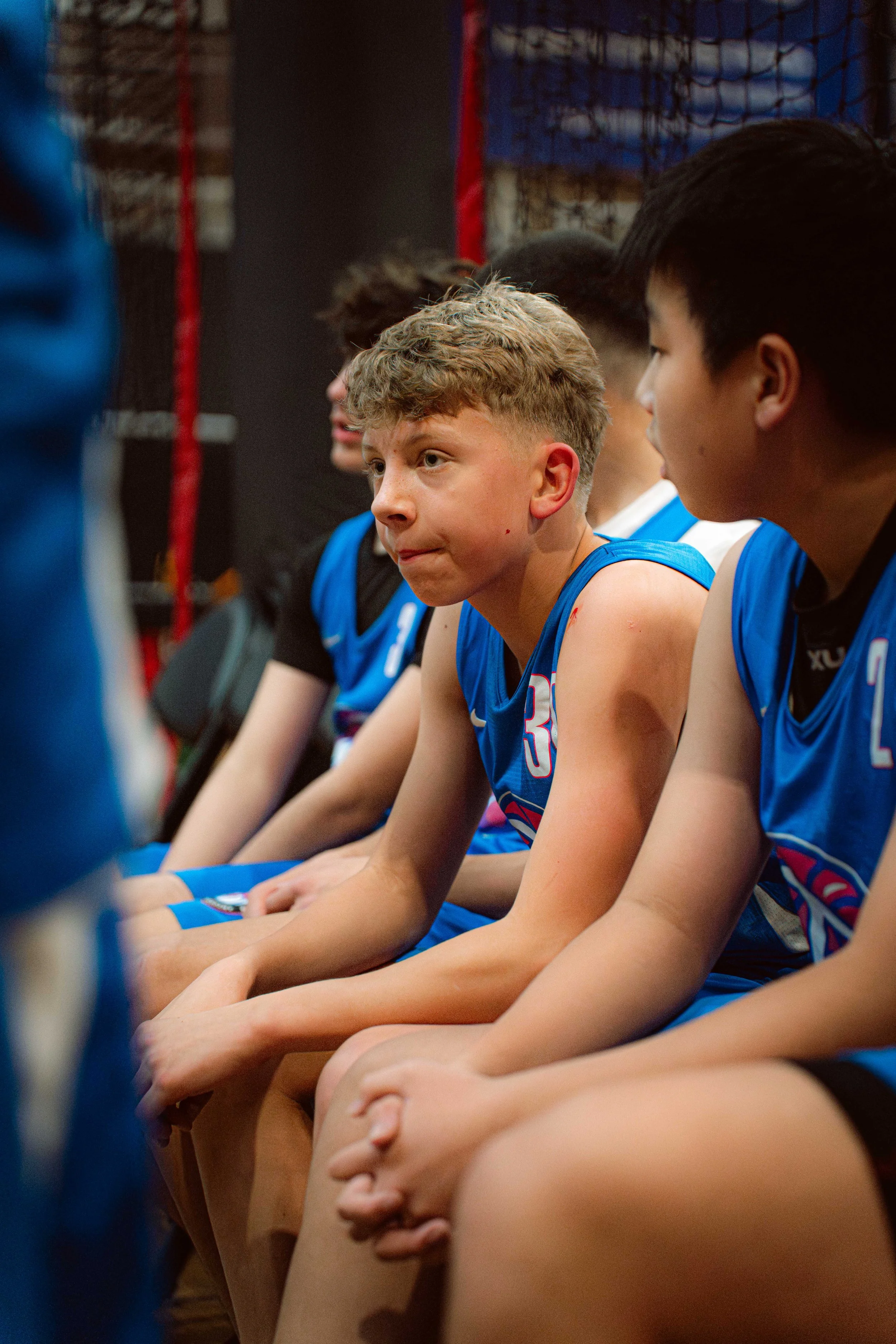 A group of young male basketball players sitting on a bench during a game or practice, wearing blue jerseys, with one player in focus looking attentive.