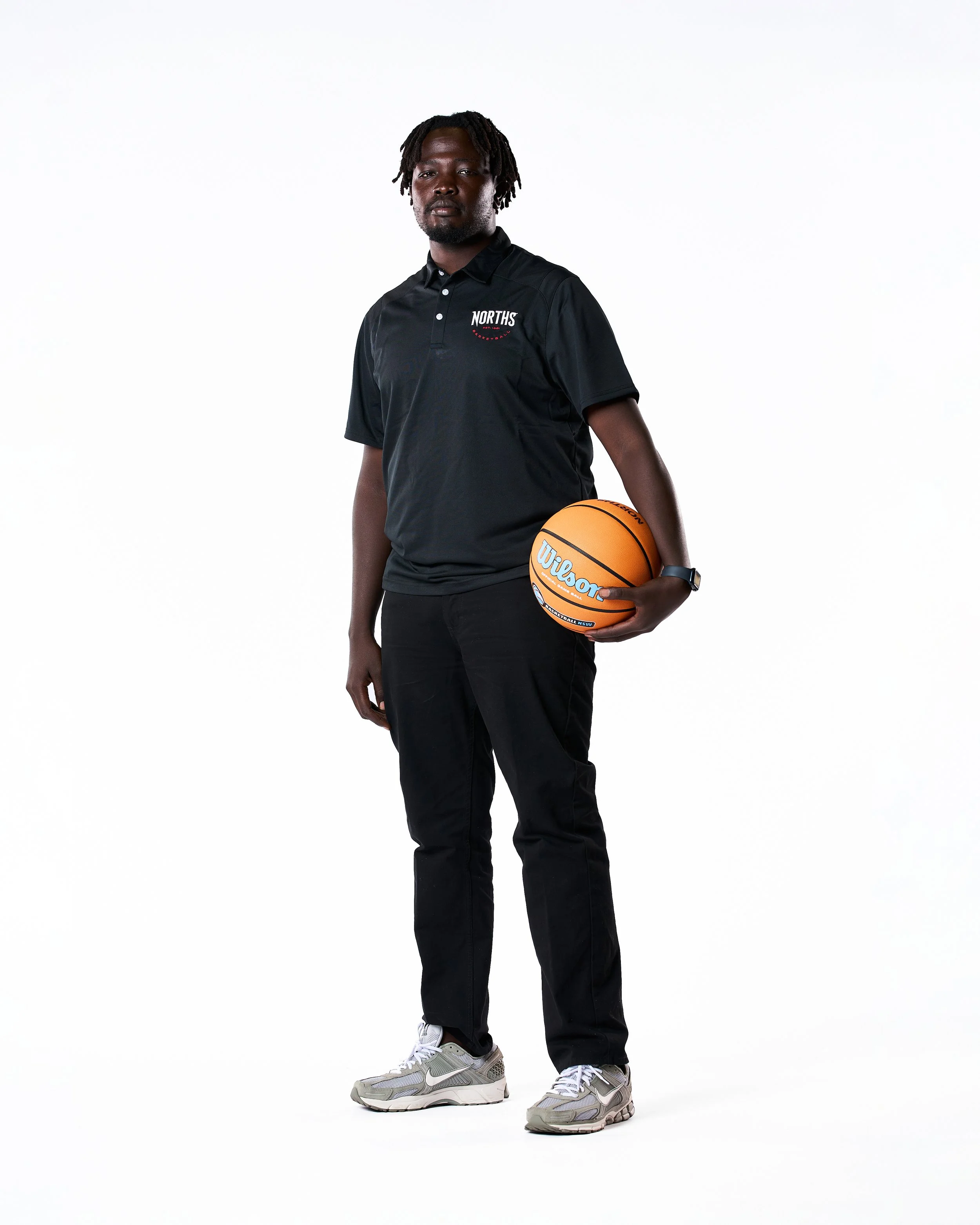 A man standing with a basketball in his right hand, wearing a black polo shirt, black pants, and gray sneakers, against a plain white background.