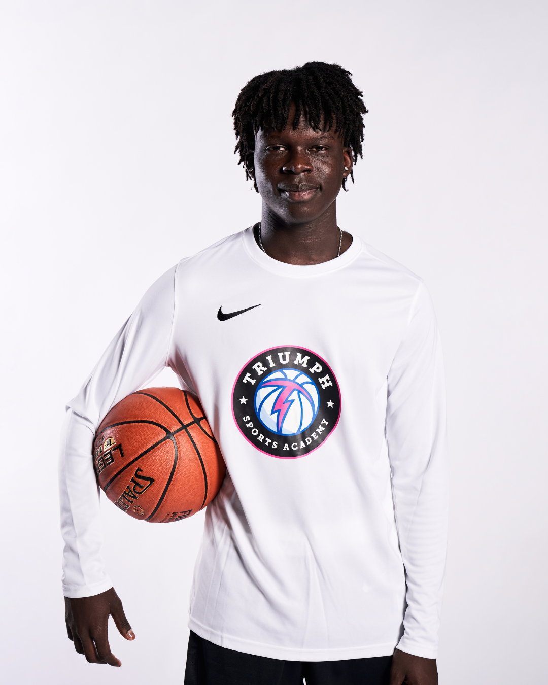 A young man with dark skin and dreadlocks holding a basketball while wearing a white sports jersey with a logo that reads 'Triumph Sports Academy' and a Nike swoosh.