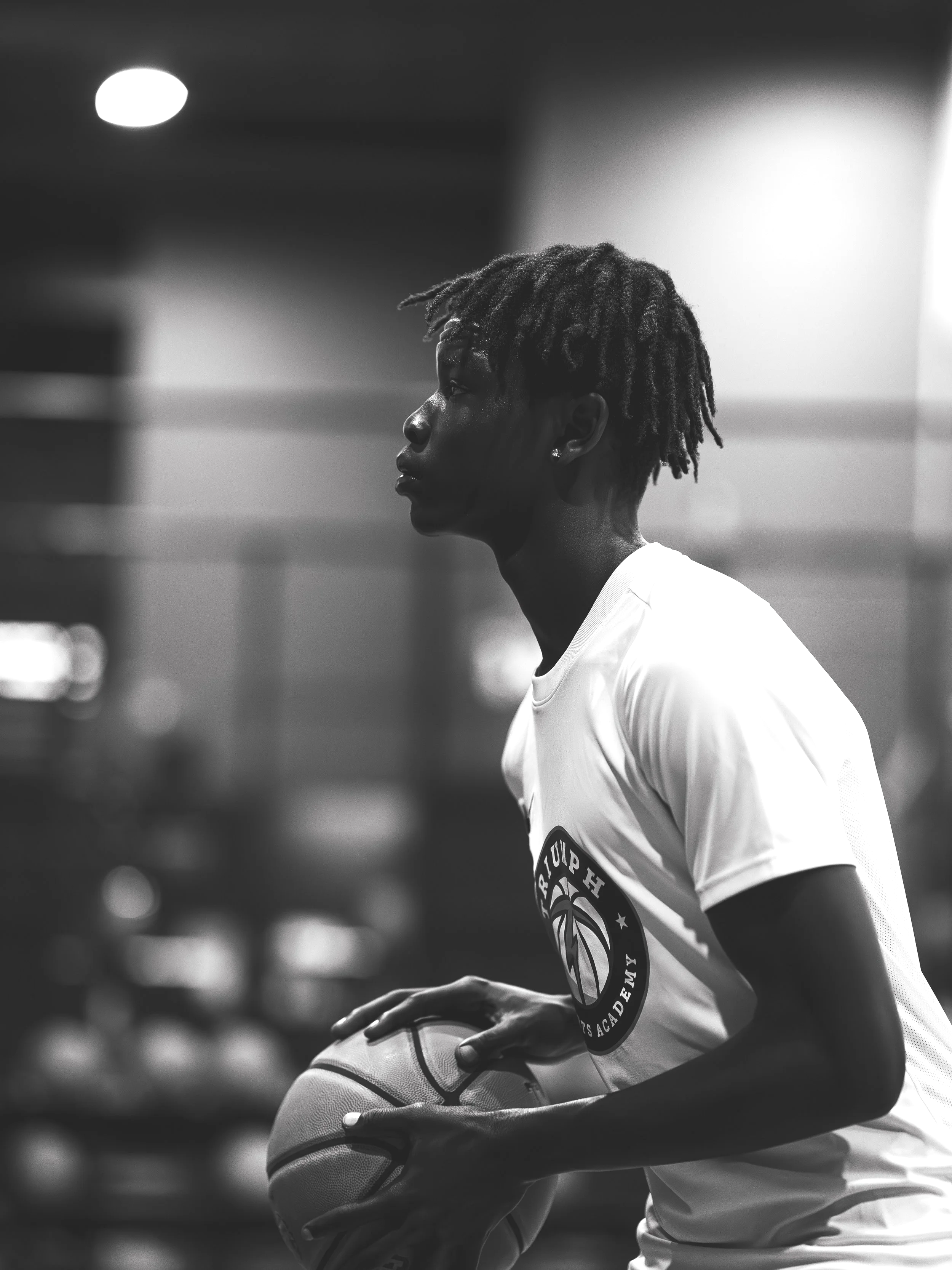 A young man with dreadlocks holding a basketball in a gymnasium, looking attentively to the side, wearing a white sports jersey with a logo on it.