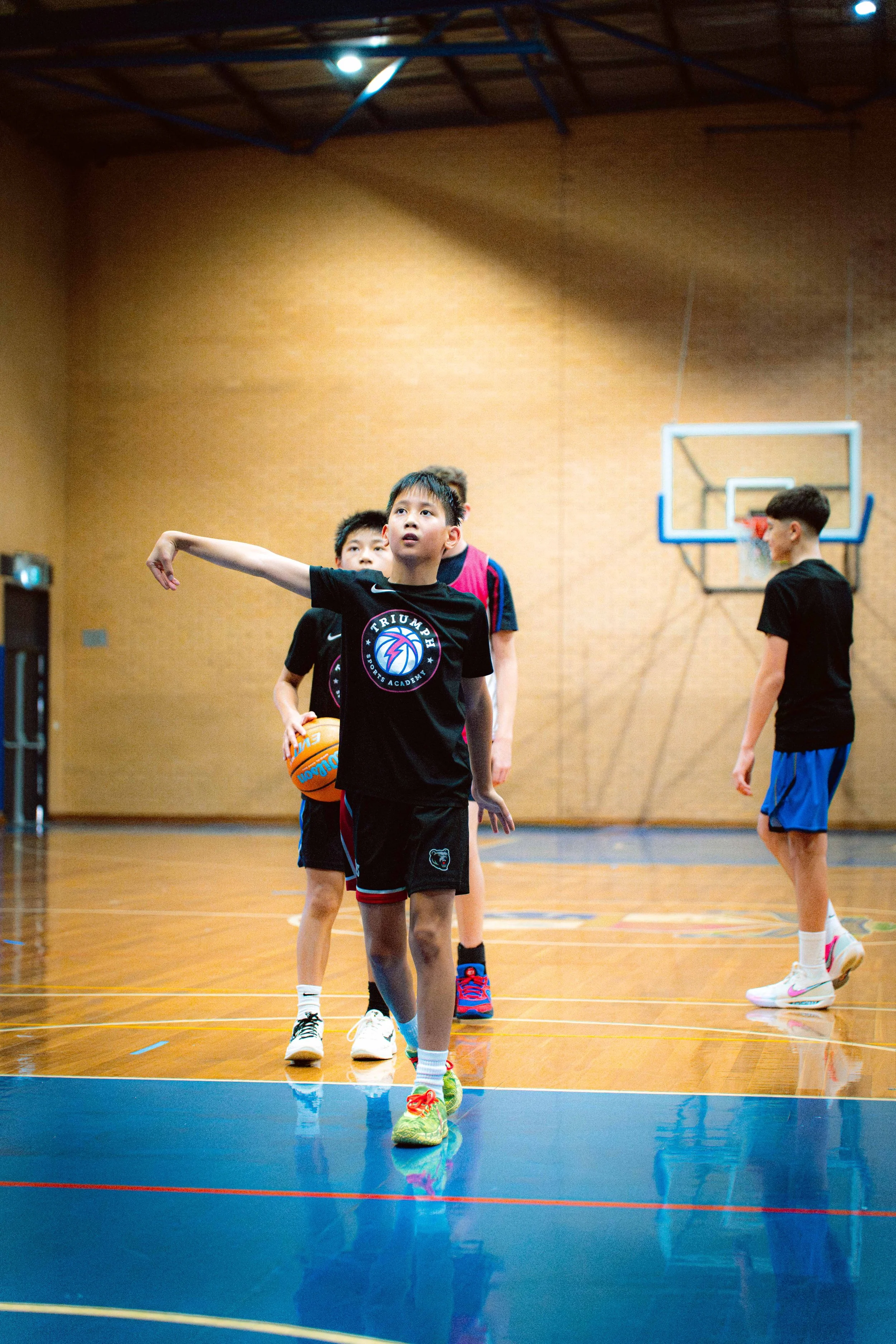 Three boys in sports attire posing for a photo on a pink backdrop. The boy in the middle is holding a basketball and wearing a pink and black jersey. The two taller boys on either side are wearing black jerseys with sports logos.