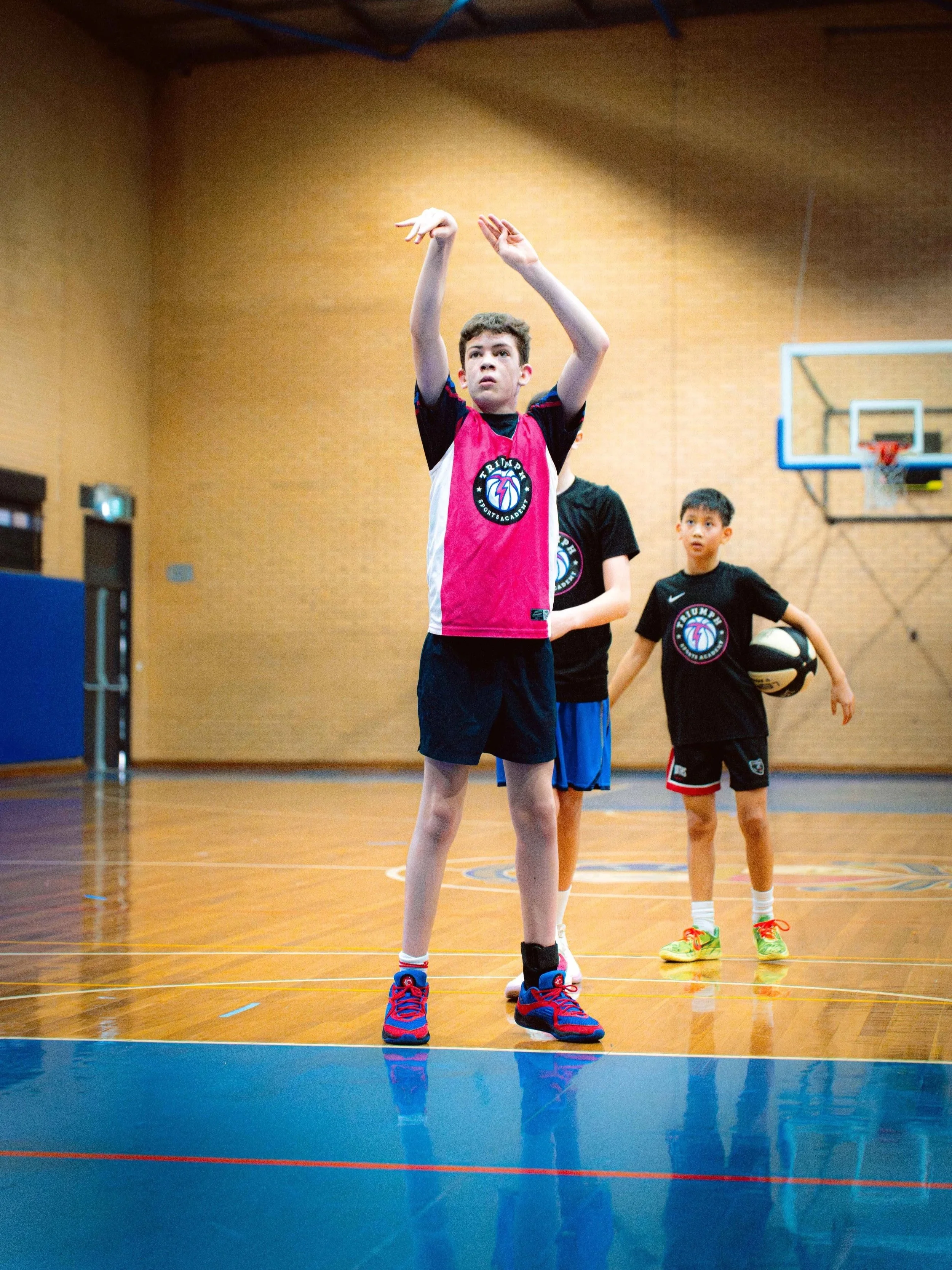 Young boys playing basketball in an indoor gymnasium with three boys visible, one actively shooting or practicing with a basketball, while the others stand nearby