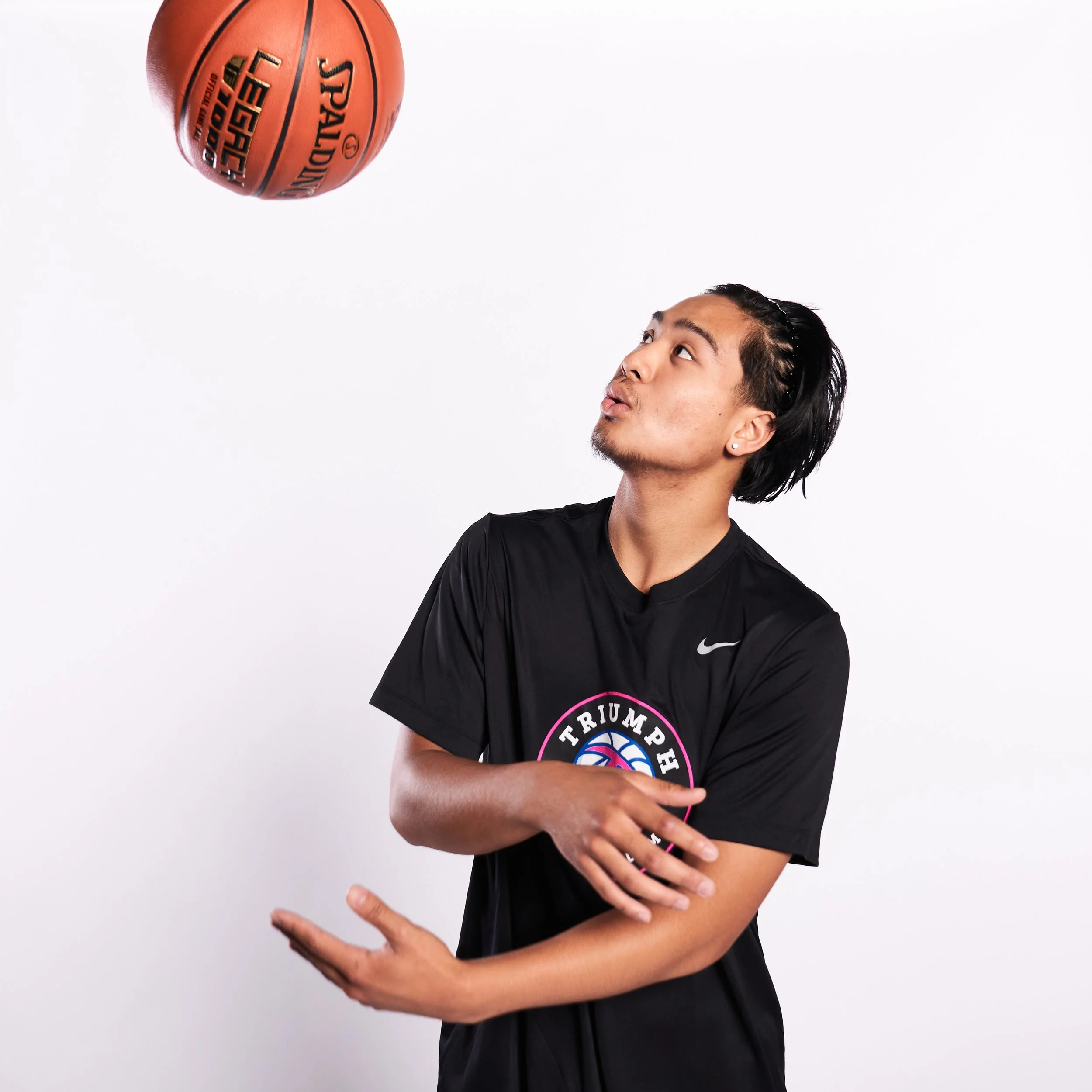 A young man in a black T-shirt with a basketball logo is preparing to catch a basketball in a studio with a plain white background.