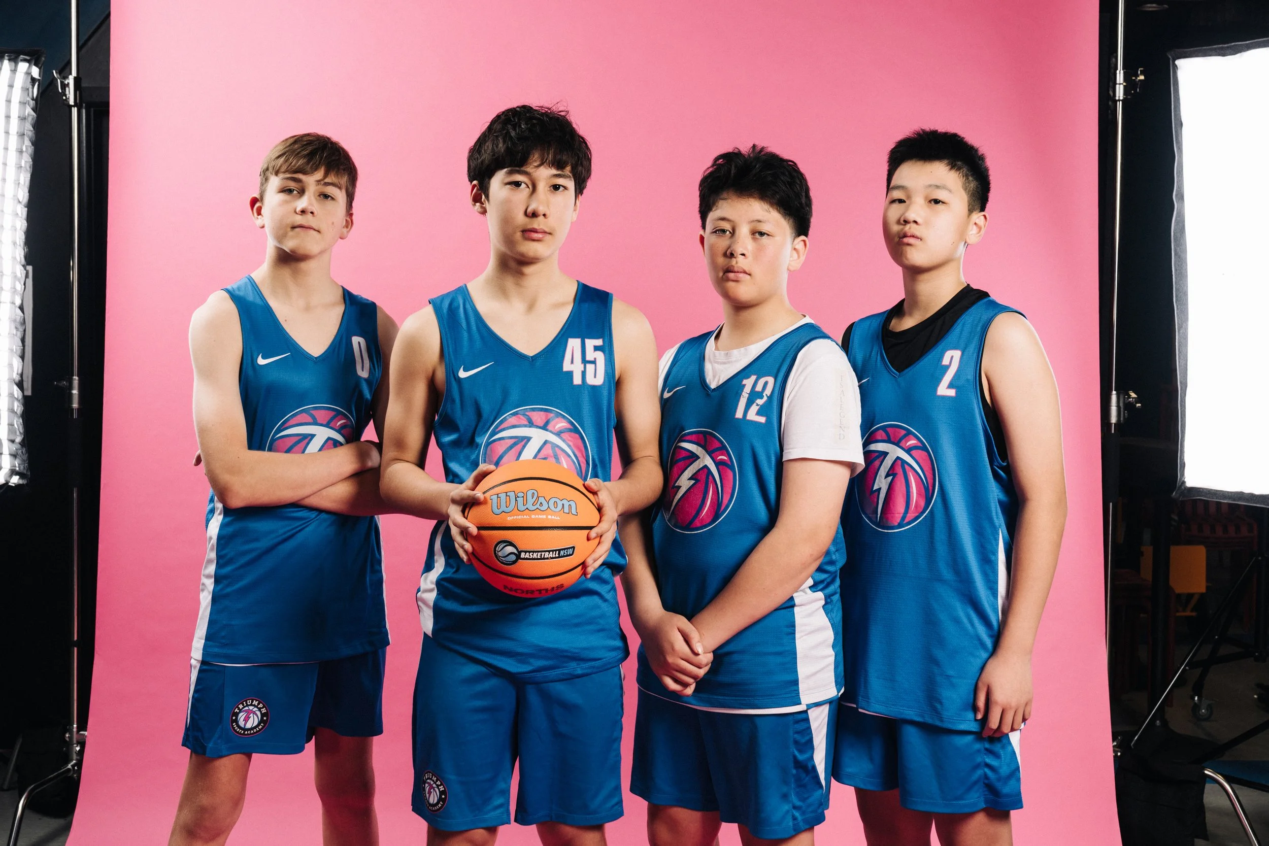 Four young boys wearing blue basketball jerseys with white accents posing in front of a pink backdrop. The boy second from the left is holding a basketball.