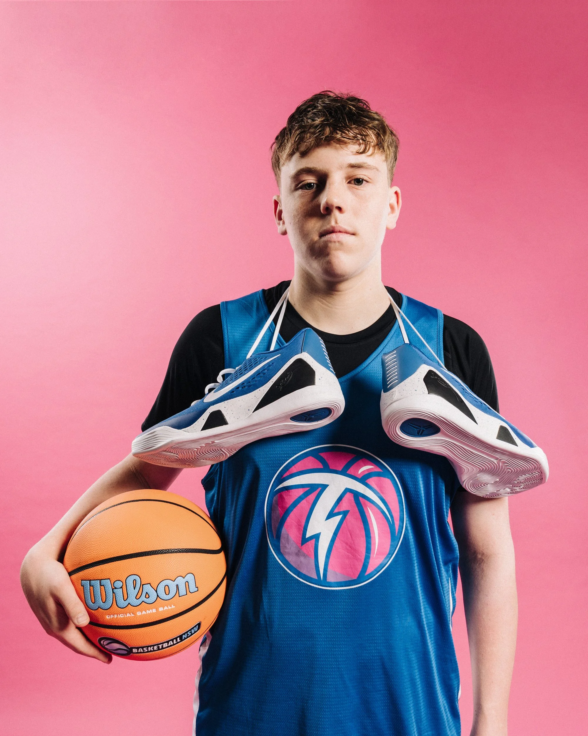 A teenage boy wearing a basketball jersey holding a Wilson basketball, with a pair of running shoes hanging around his neck, standing against a pink background.