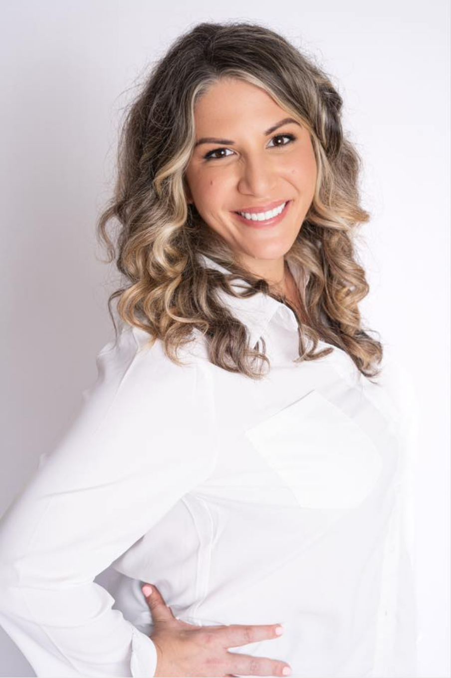 Portrait of a smiling woman with wavy blonde hair wearing a white shirt, standing against a plain light background.