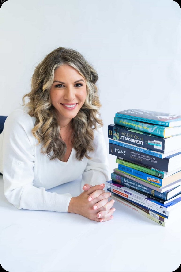 A woman with blonde curly hair, smiling and sitting at a white table, next to a tall stack of books on mental health and therapy.
