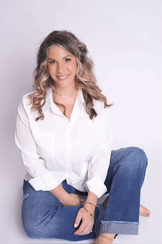 A young woman with wavy blonde hair sitting on the floor against a plain white background, wearing a white button-up shirt and blue jeans, smiling at the camera.