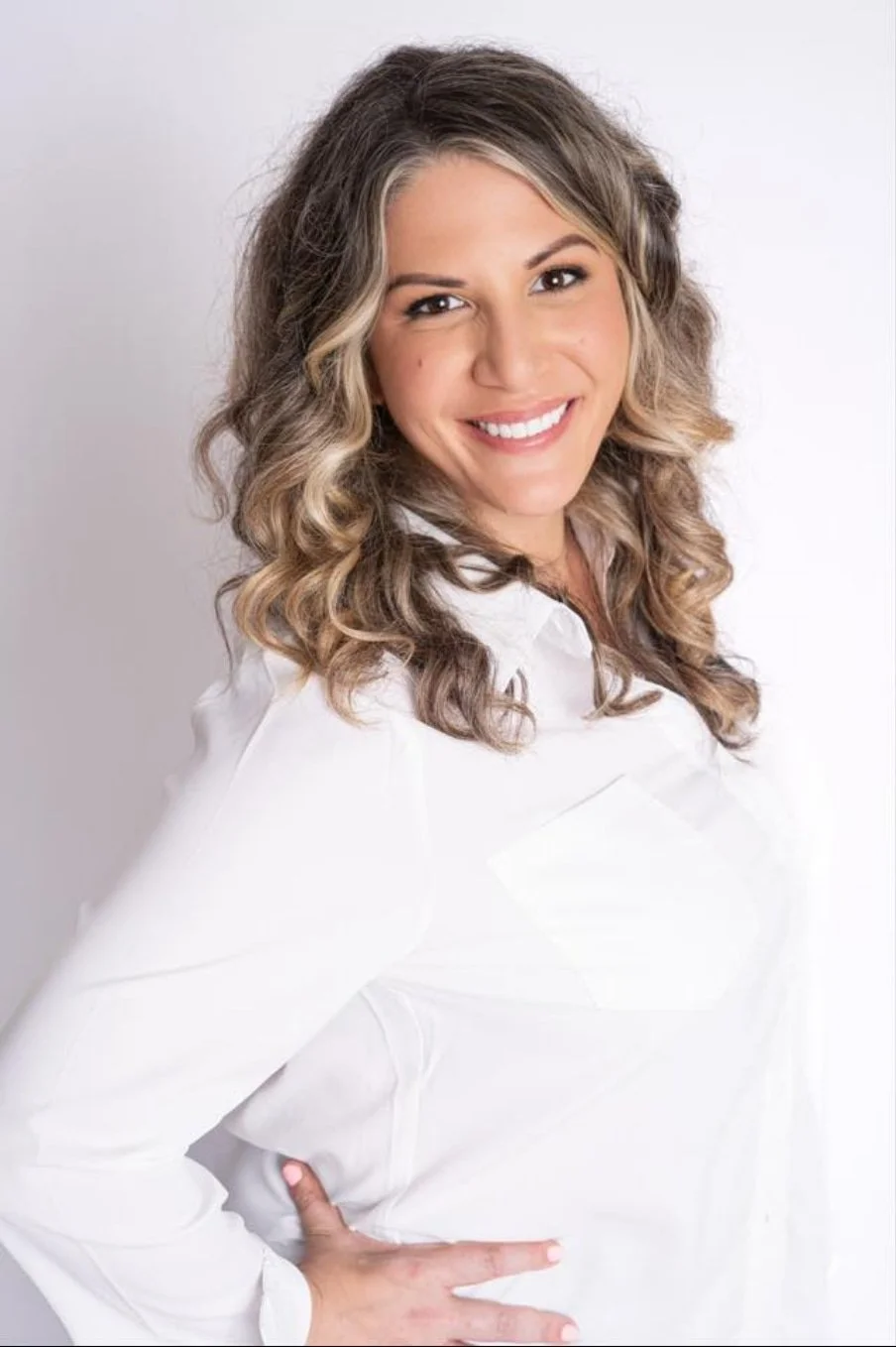 A smiling woman with wavy hair wearing a white shirt stands against a white background, hand on her hip.