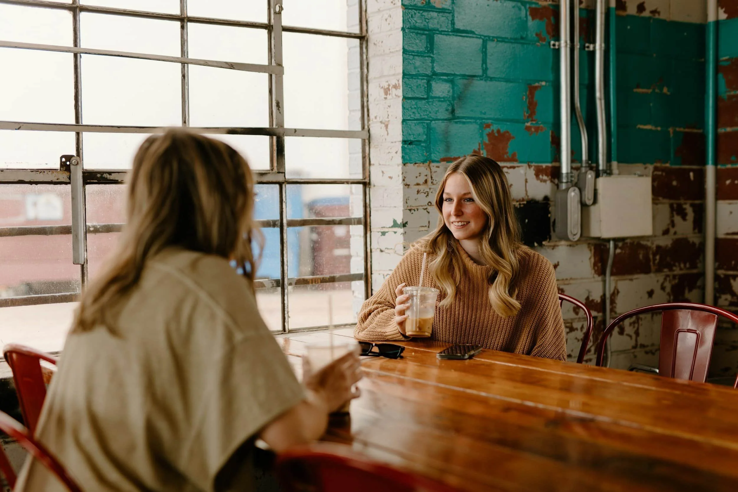 Two women sit at a wooden table in a café, chatting and smiling while holding iced drinks by a large window.