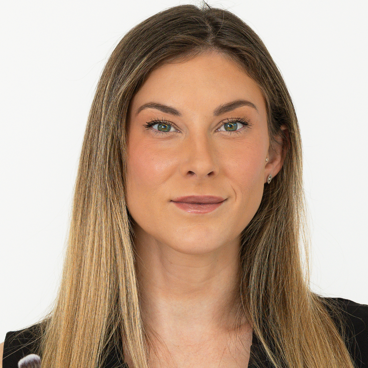 A woman with long, straight, light brown hair, fair skin, green eyes, and a neutral expression, wearing a black top and small earrings, against a plain white background.
